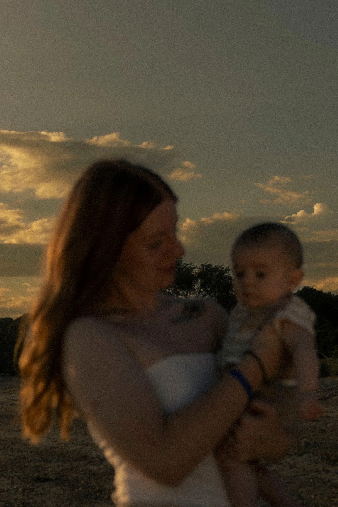 A woman holding a baby during sunset with a cloudy sky and trees in the background.
