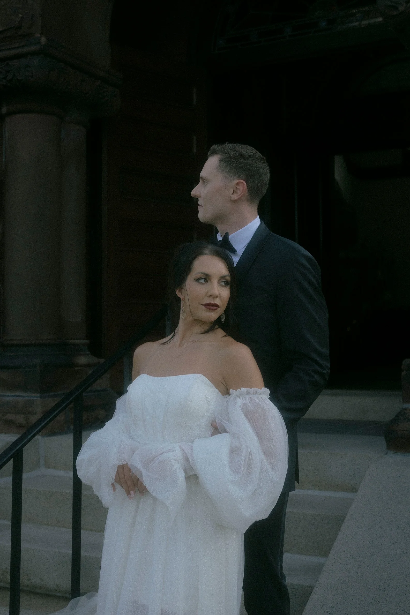 A man and woman in wedding attire are standing outside a church, with the woman in a white wedding gown and the man in a black tuxedo.