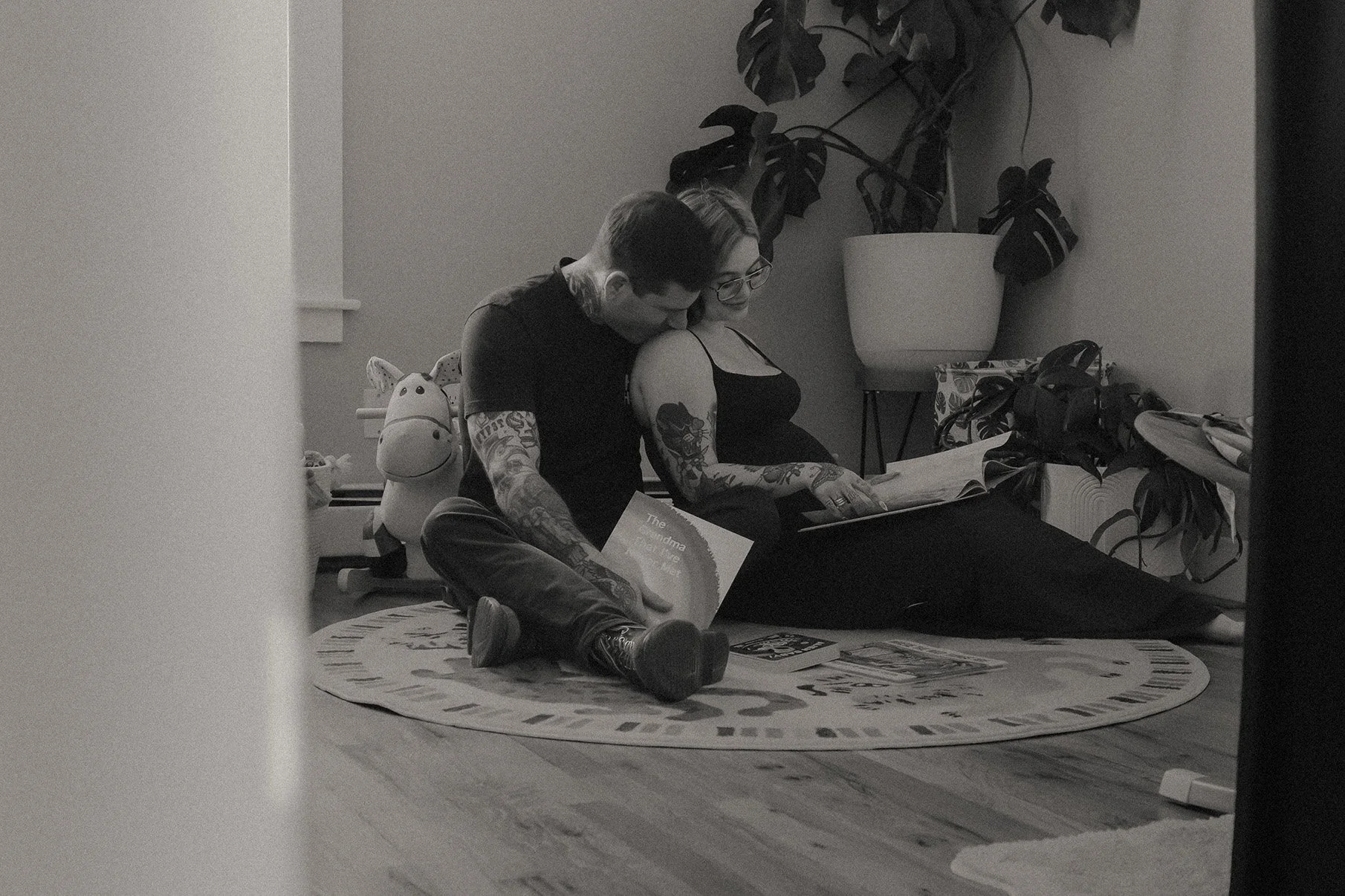 A couple sitting on a round rug inside a room, reading a book together with a large potted plant nearby. The man has tattoos and the woman has glasses and tattoos on her arms. There are children's toys and books around them.