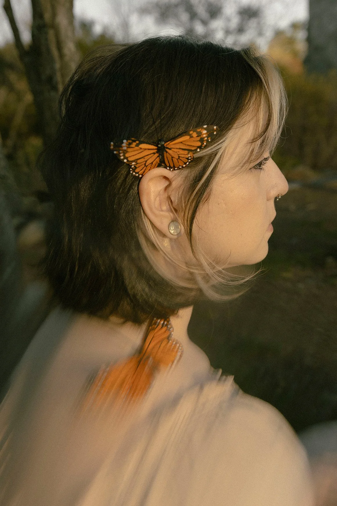 A woman with short brown hair featuring blonde highlights, wearing a Monarch butterfly as a hair accessory, plug tunnels, with a matching butterfly sits on her shoulder, outdoors in a natural setting. There is a motion blur at the bottom of the image
