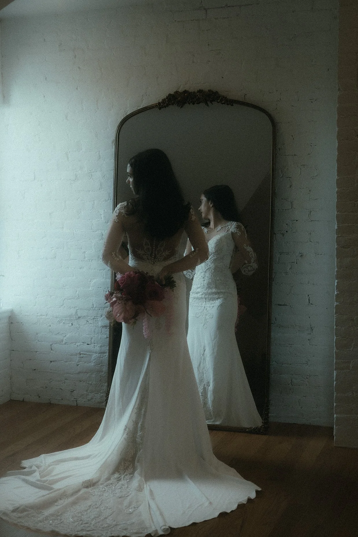 Bride in a lace wedding dress holding a bouquet stands in front of a large mirror, her reflection shows her smiling, against a white brick wall in a dimly lit room.
