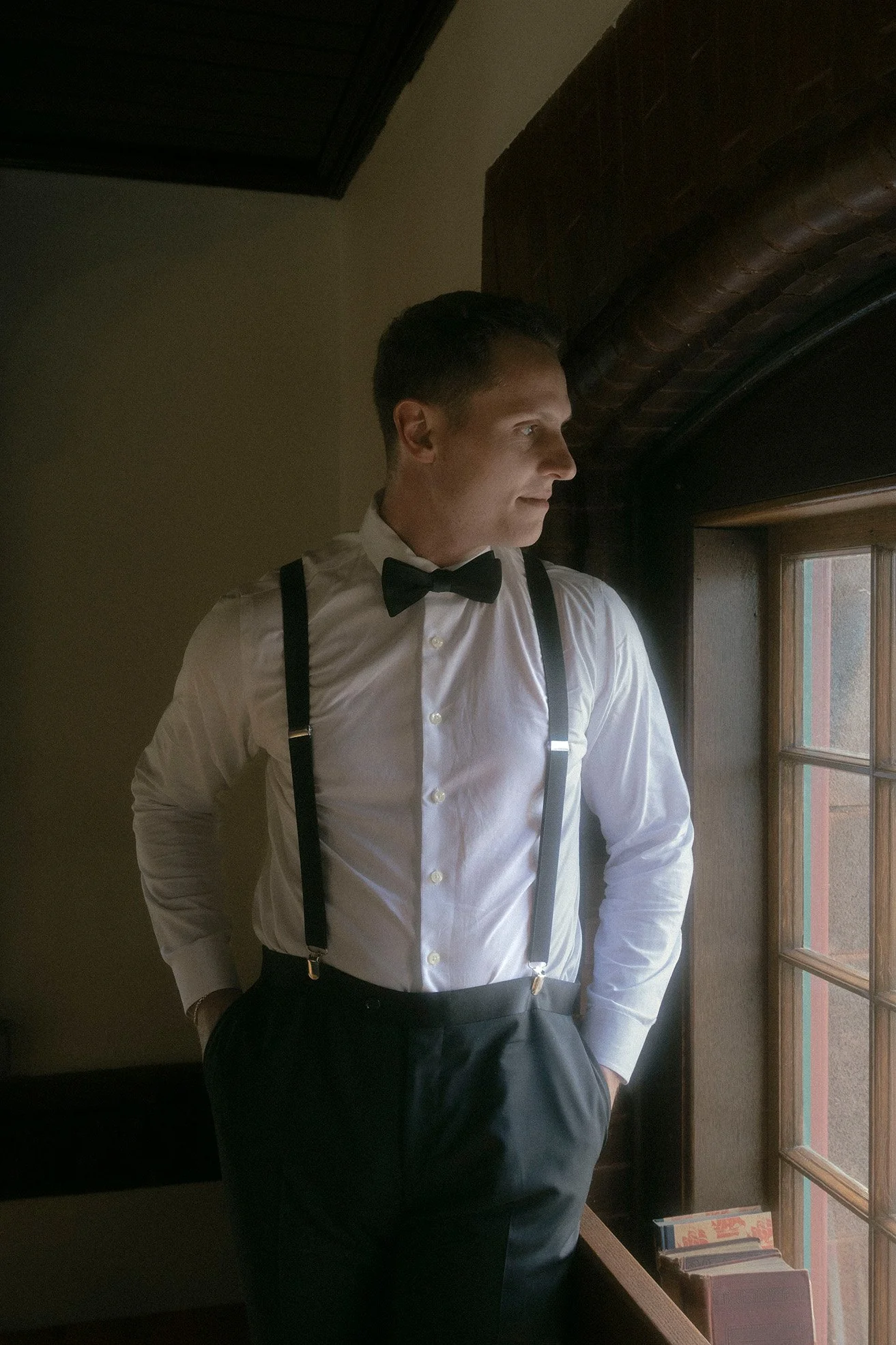Man (groom) wearing a white dress shirt, black bow tie, and suspenders standing by an old window in a historical building and gazing outside.