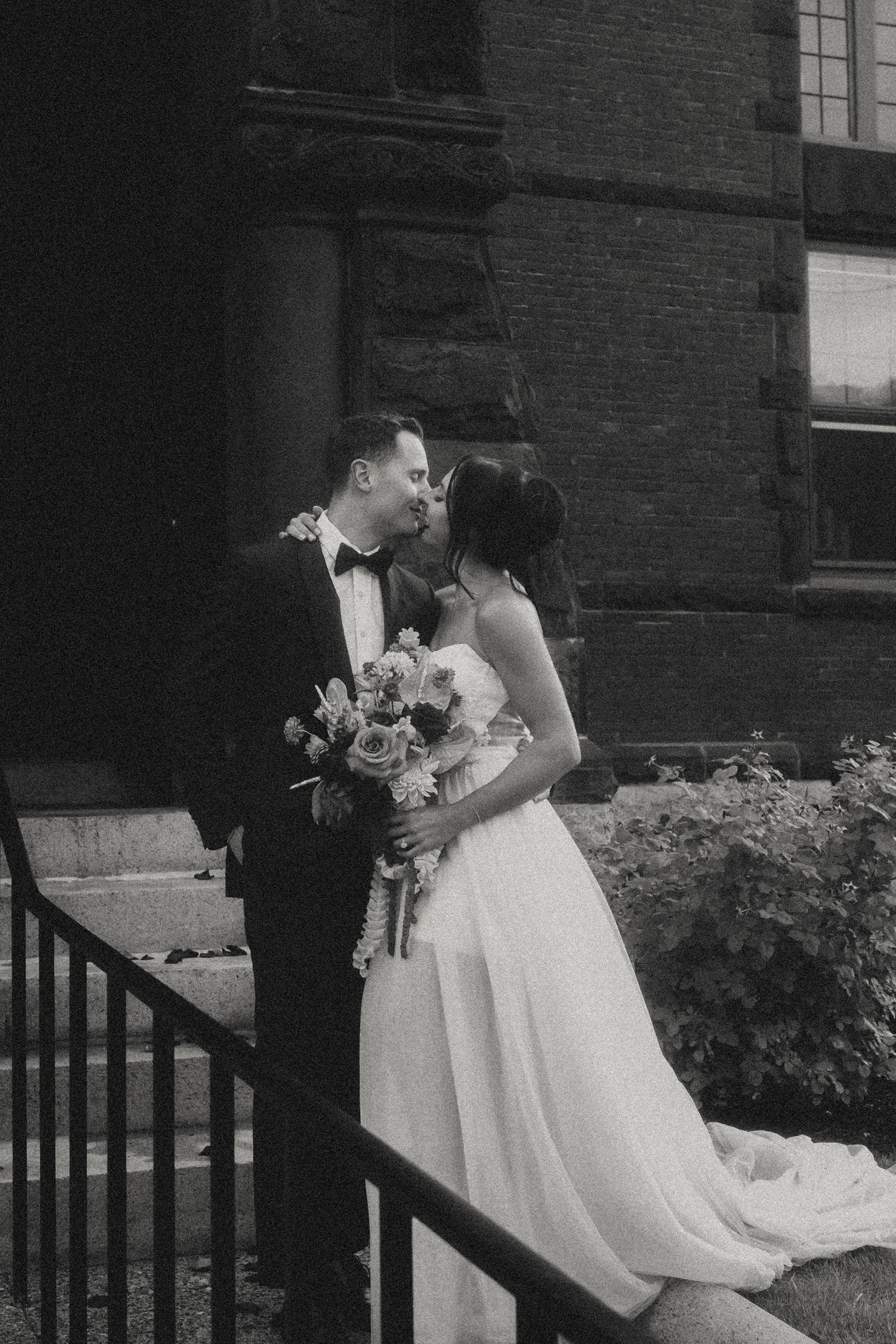 A wedding couple sharing a kiss outdoors, with the groom in a tuxedo and the bride in a white wedding gown holding a bouquet of flowers.