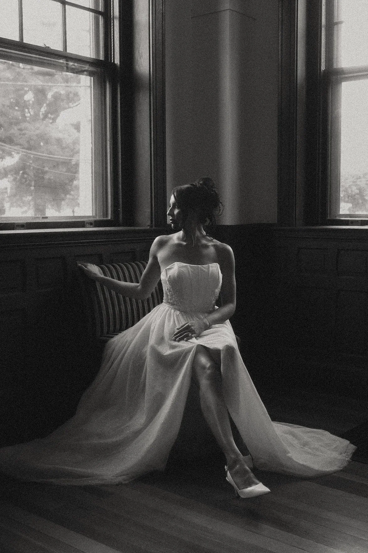 A classic, timeless black and white photograph of a woman in a sleeveless wedding dress sitting on a chair gazing out the window in a dimly lit room.