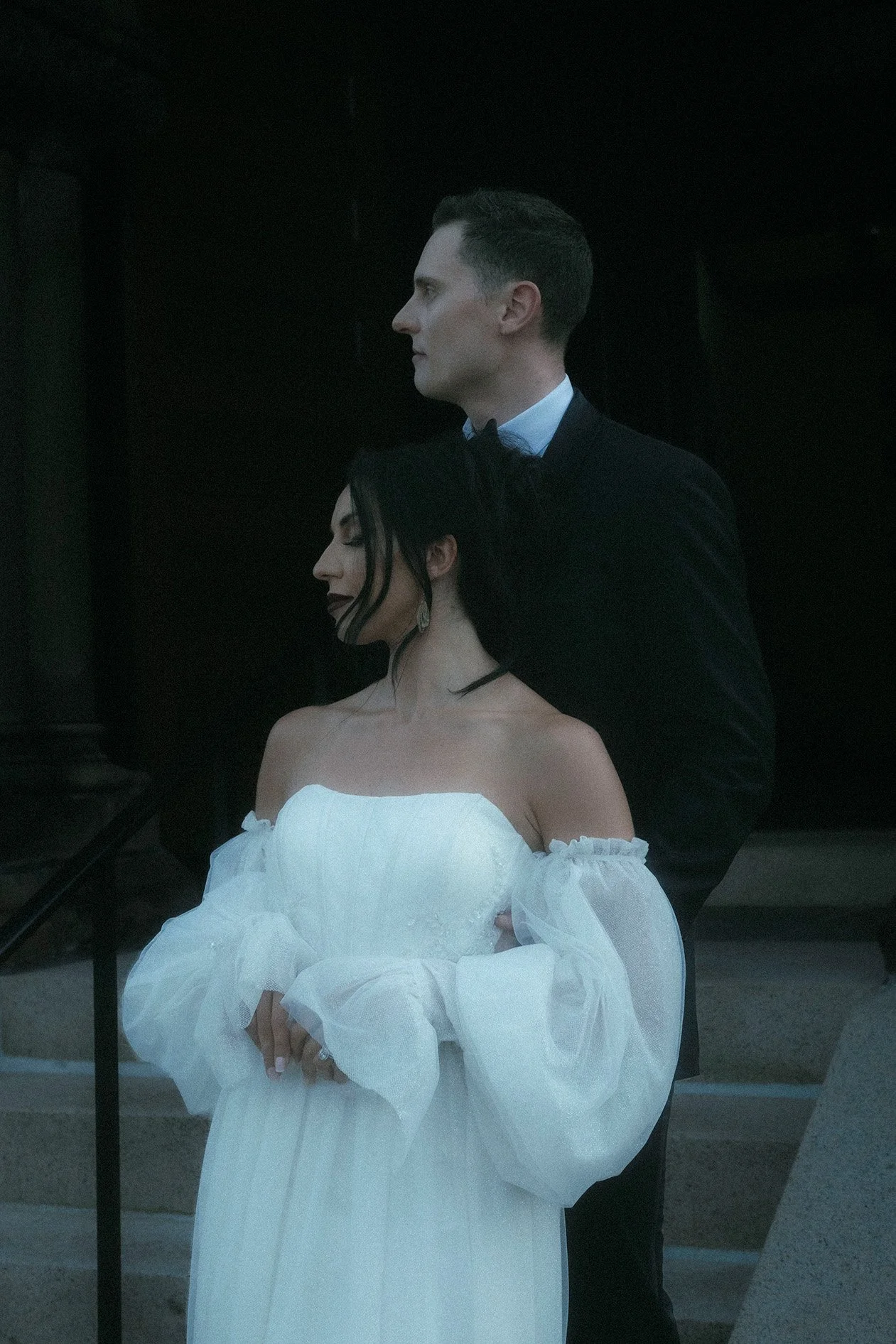 A man in a black tuxedo and a woman in a white off-shoulder dress standing together, with the woman slightly in front of the man, against a dark background.