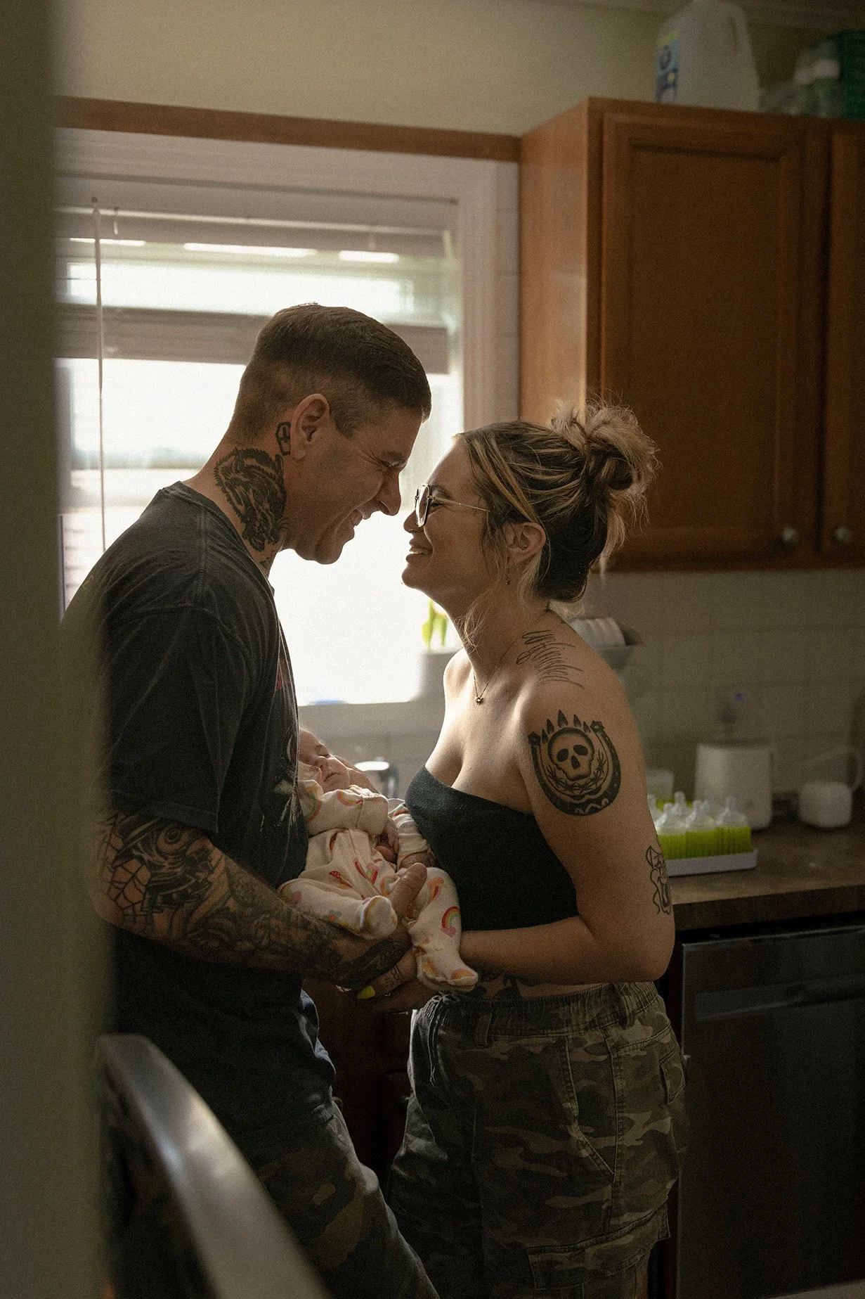A couple holding a newborn baby in a kitchen, smiling at each other. The man has tattoos on his arm and neck, and the woman has tattoos on her shoulder and wears glasses. They are standing close near a window with blinds.