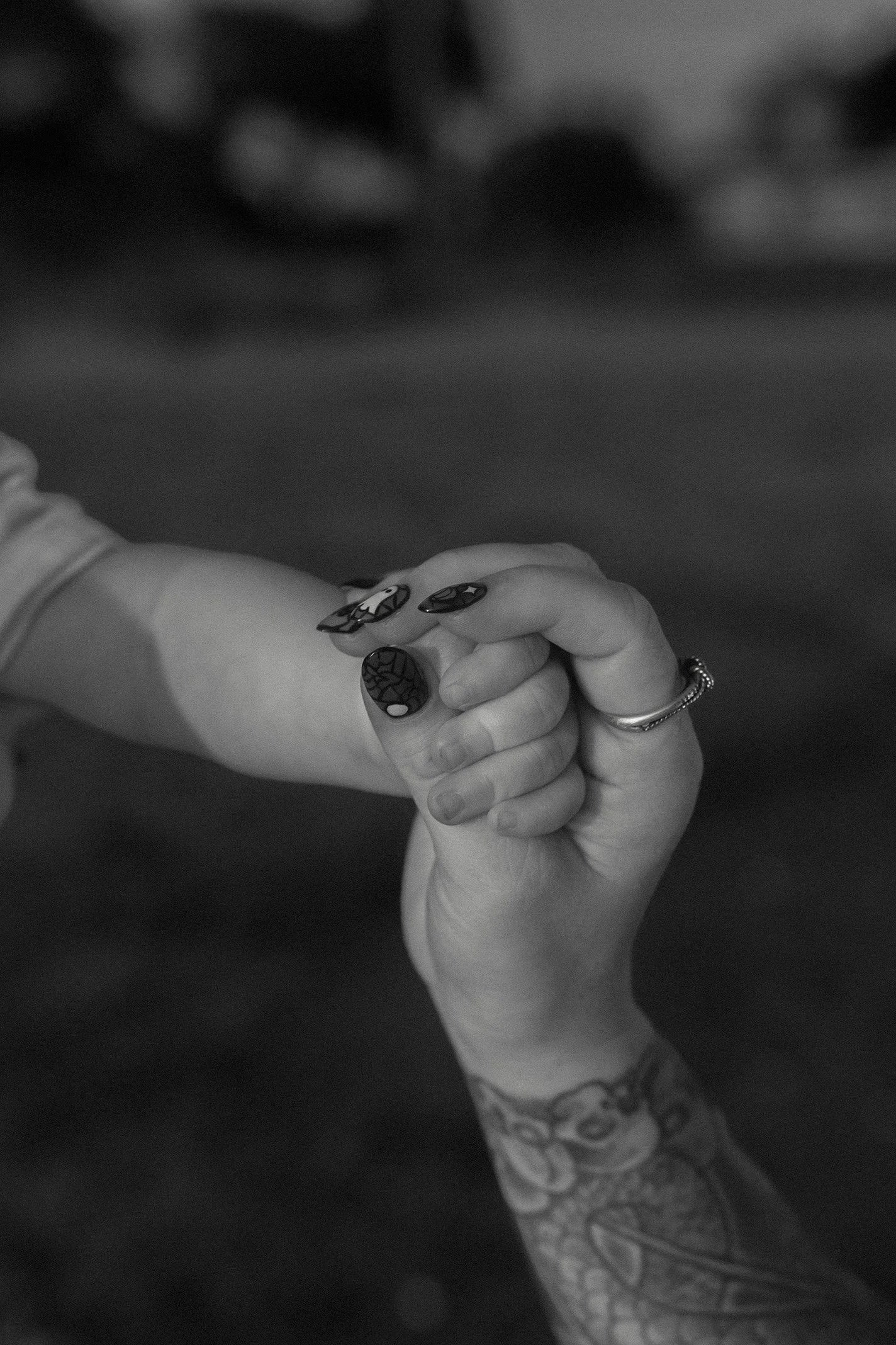 A person with a tattoo on their forearm holding a child's hand, both with interlocked fingers, black nail polish with designs, and a ring on the person's finger.