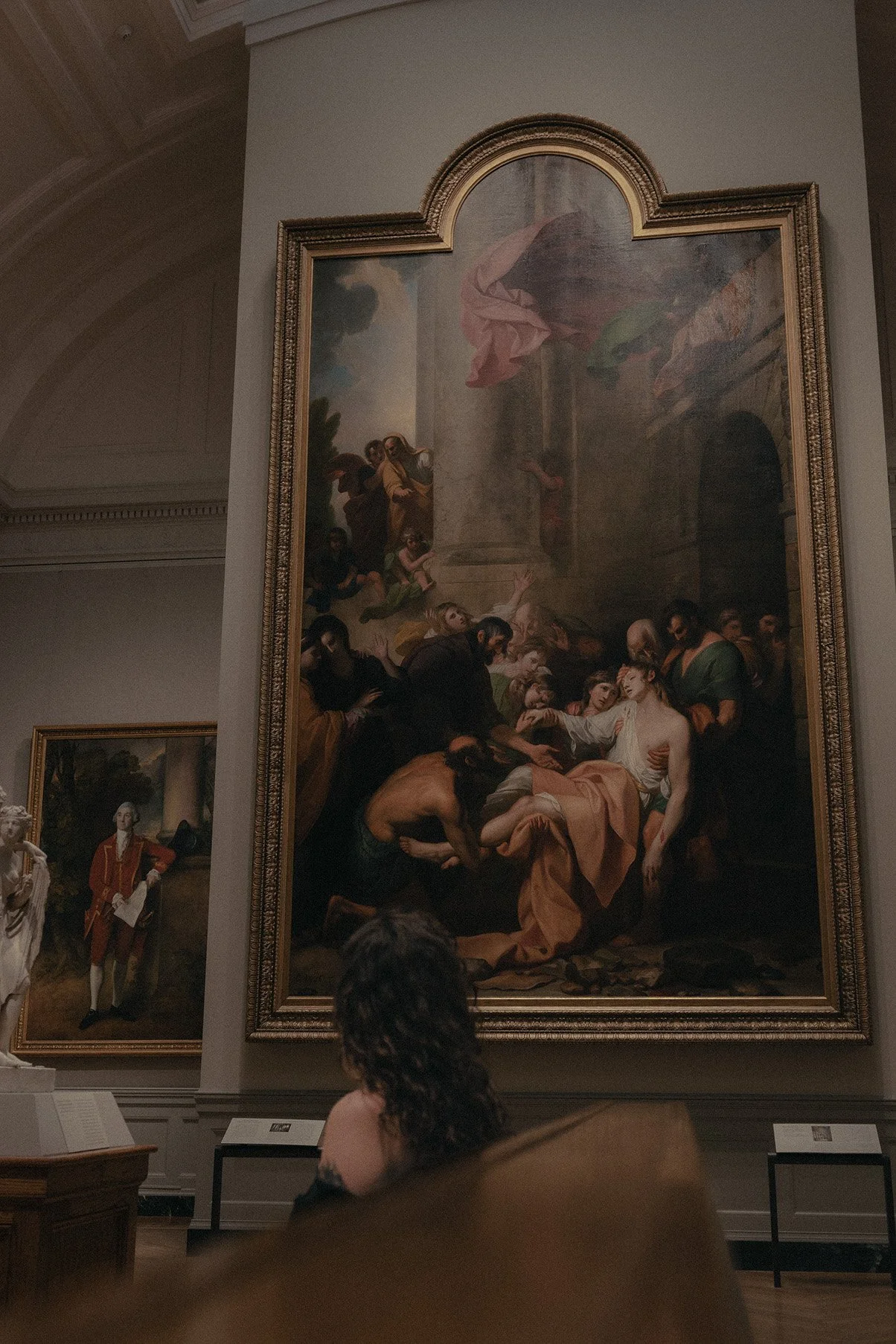 A woman with curly hair sitting in front of a large religious artwork in a museum or gallery.