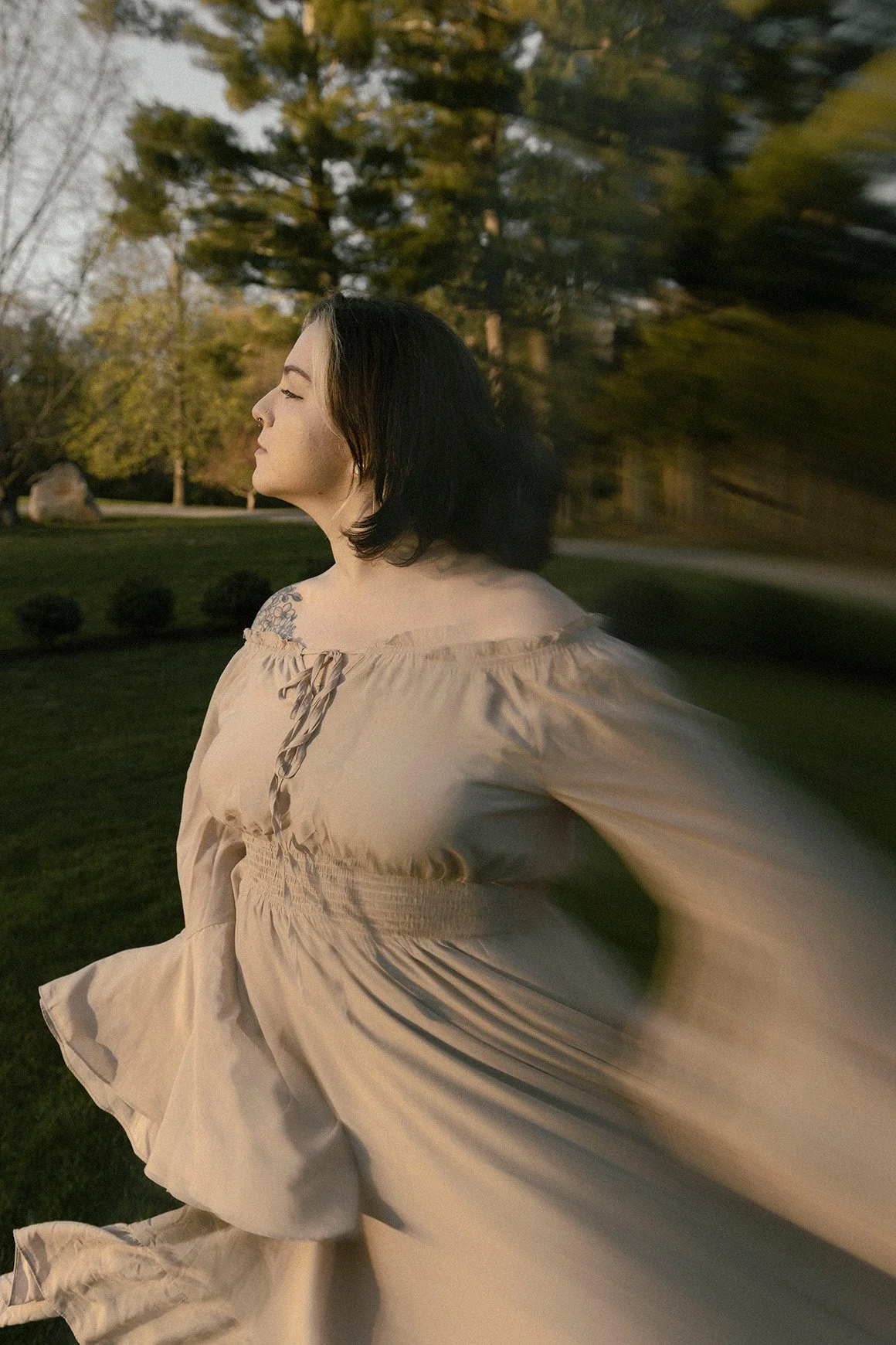 A woman with short dark hair and a tattoo on her left shoulder, standing outdoors in a park with trees and a lawn, dressed in a flowing beige dress, with her eyes closed and her face turned slightly to the left, captured in a motion-blurred photograp