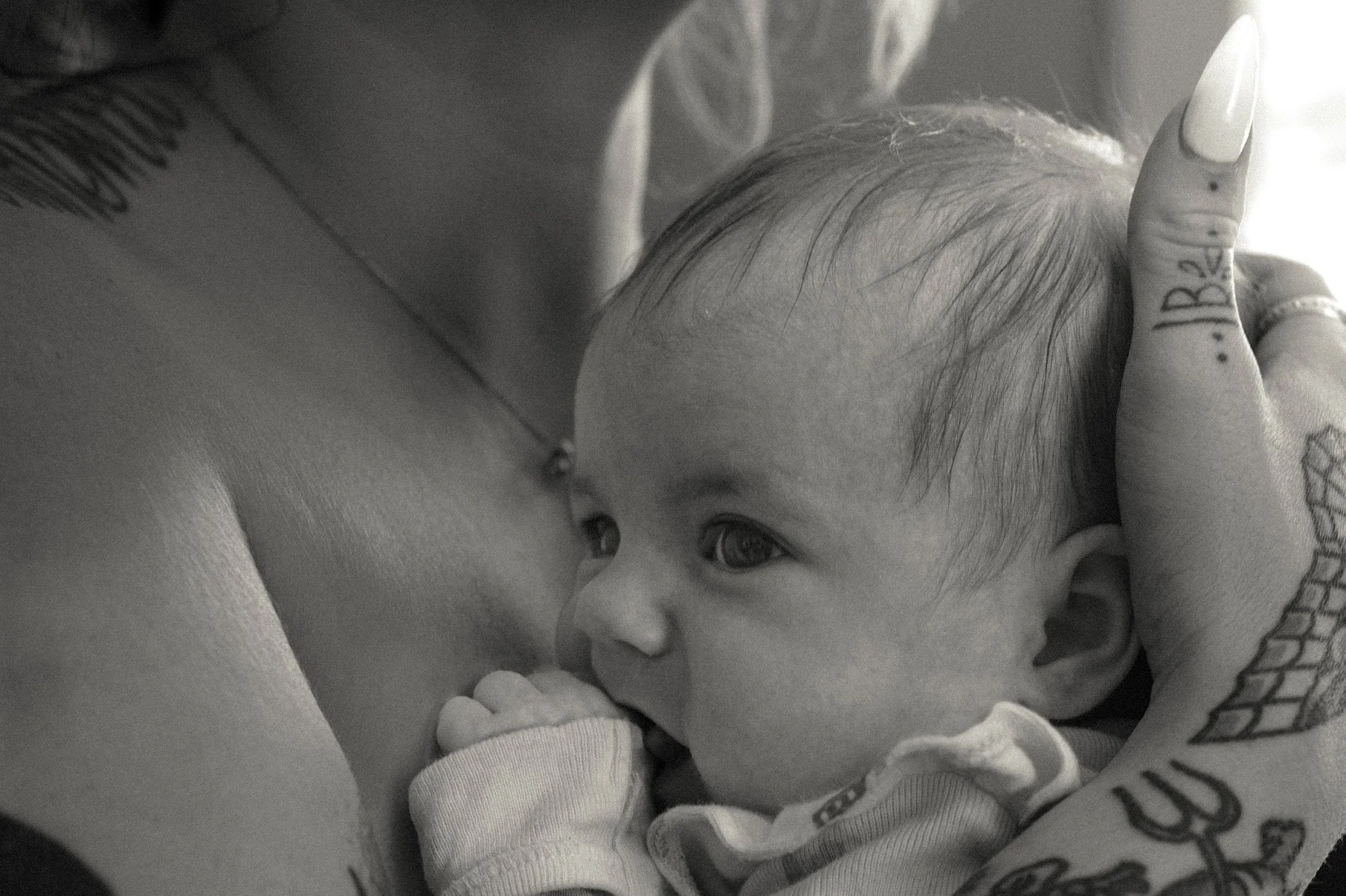 A close up image of a baby girl held close to her mother's bare chest, her first to her mouth as she looks off to the side. Her mother has long nails with tattoos visible on the hand.