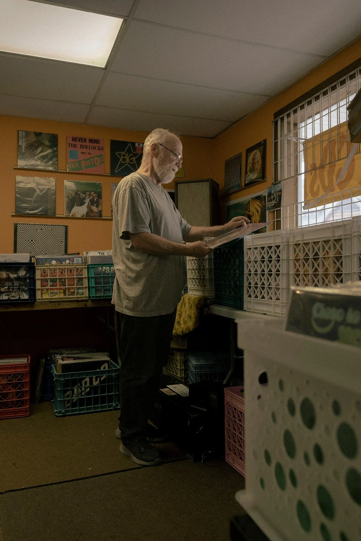 An older man with glasses and a gray t-shirt browsing through records or magazines in a small record store, with vinyl album covers displayed on the walls behind him.