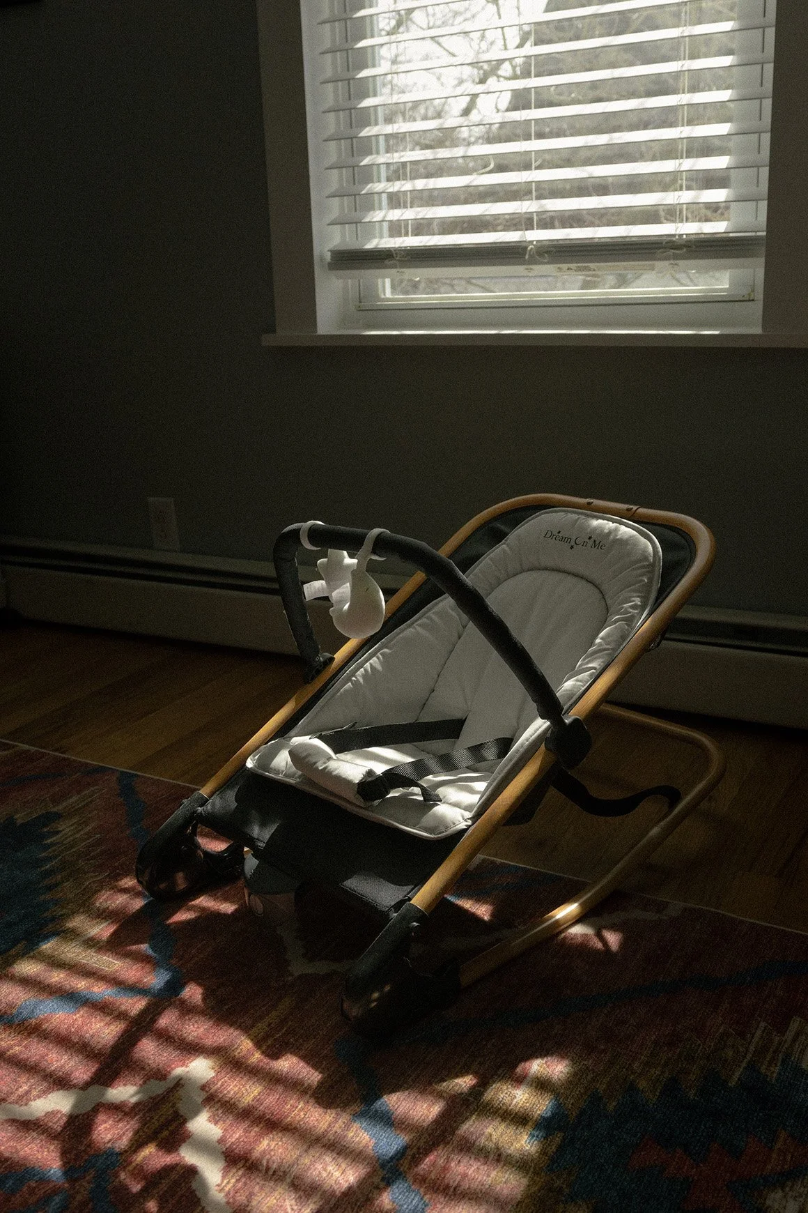 A baby bouncer with a white cushioned seat and wooden frame placed on a patterned rug in front of a window with blinds.