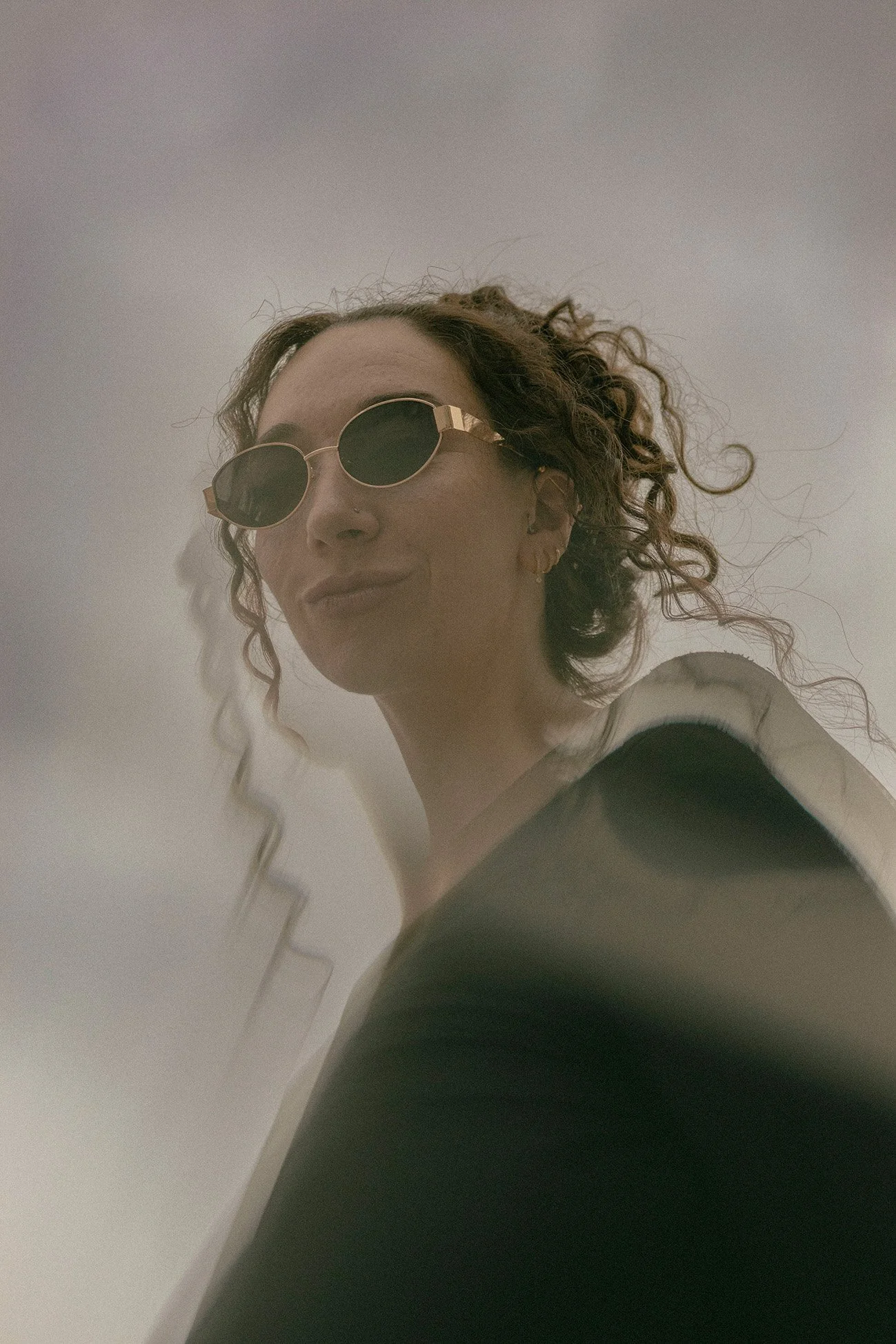An angled-up photo of a woman with curly hair wearing round sunglasses and a black top, smiling slightly, with a grey sky background.