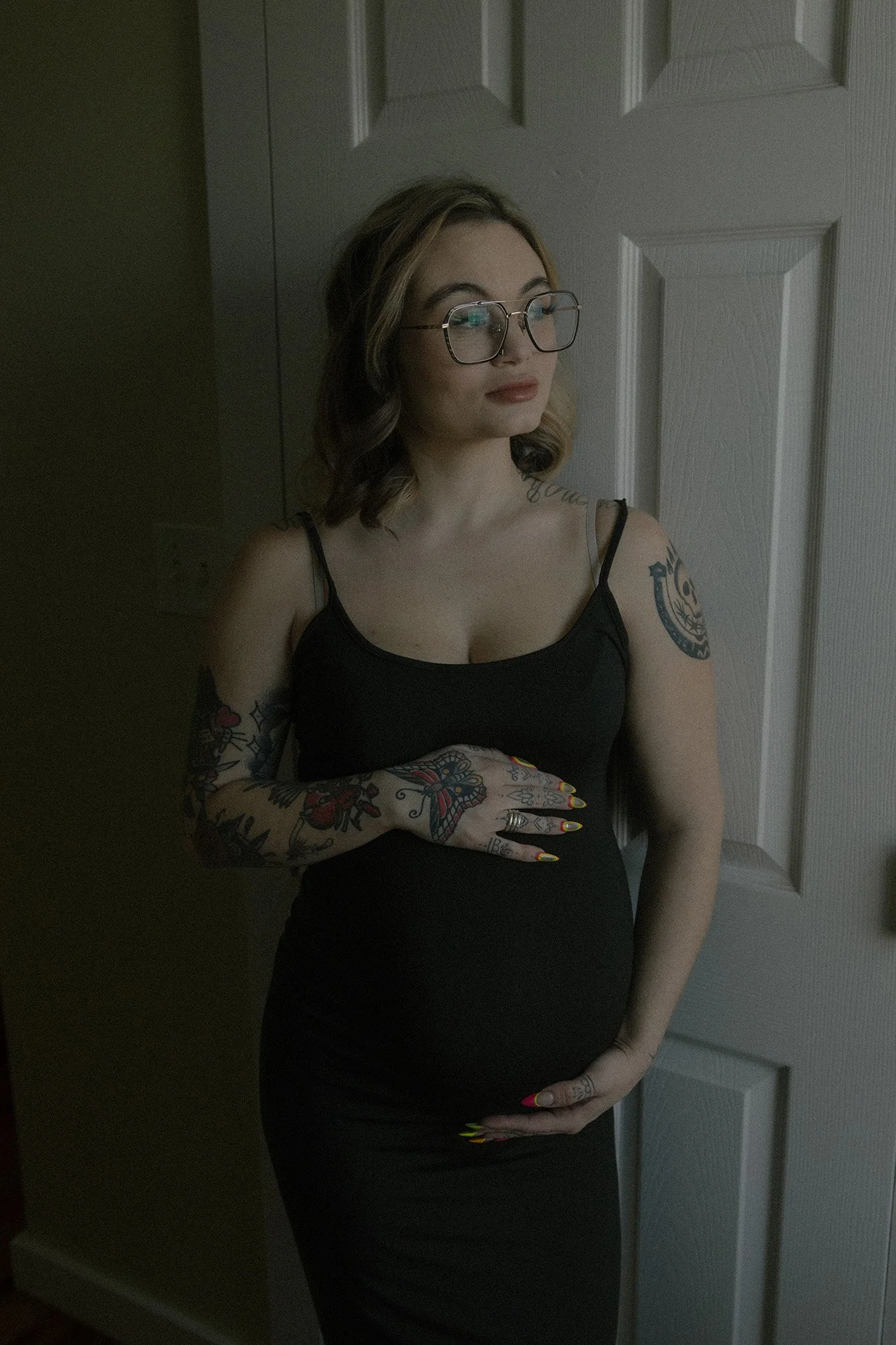 A woman with tattoos wearing glasses and a black dress, standing in front of a white door, as she gently caresses her pregnant belly.