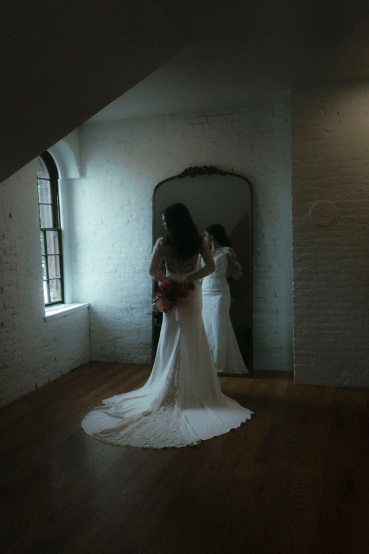 A bride in a lace wedding dress standing in front of a mirror, holding a bouquet of flowers, in a room with white brick walls and wooden floor, lit by natural light from a window.