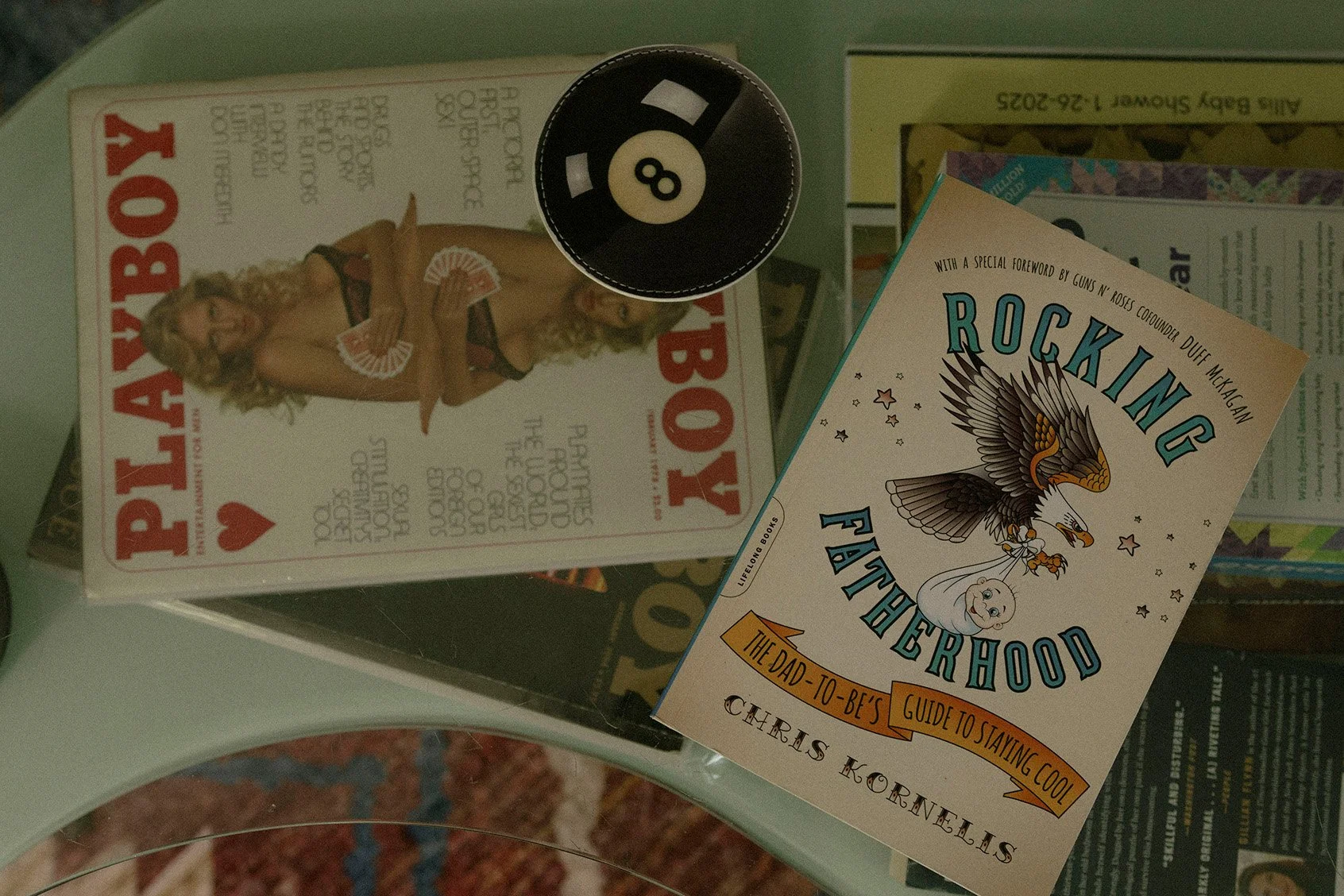 A magazine titled "Playboy" featuring a woman in lingerie holding playing cards, a black 8-ball, and a paperback book titled "Rocking Fatherhood" on a glass surface.