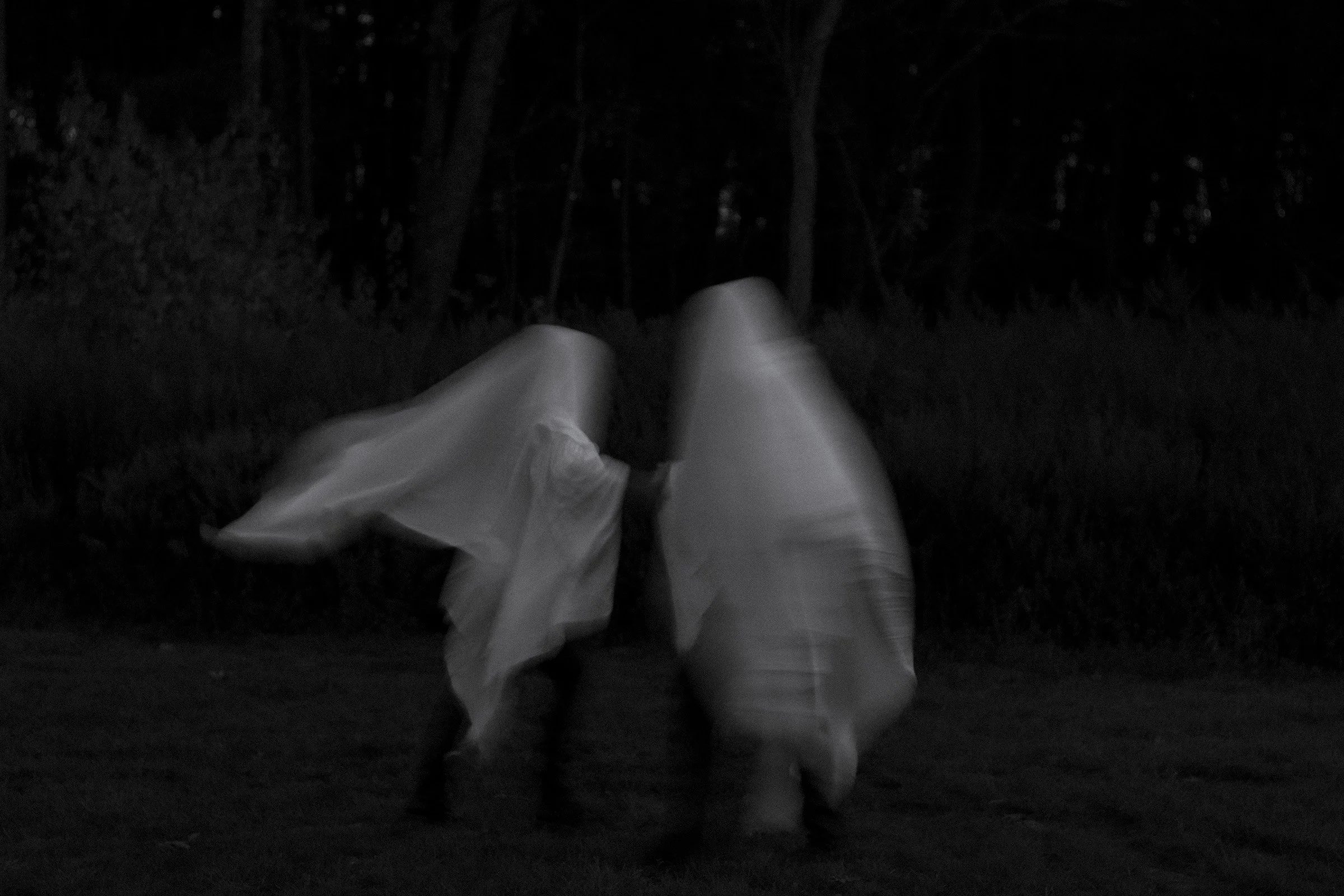 Two people wearing flowing white garments walking through a dark forested area at night.