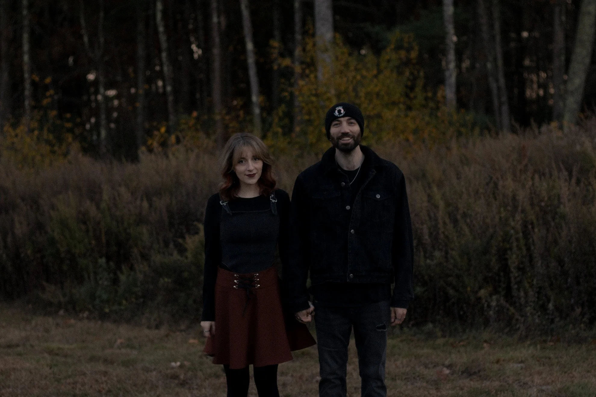 A young woman and a man holding hands, standing outdoors in a forested area with trees and autumn foliage in the background.