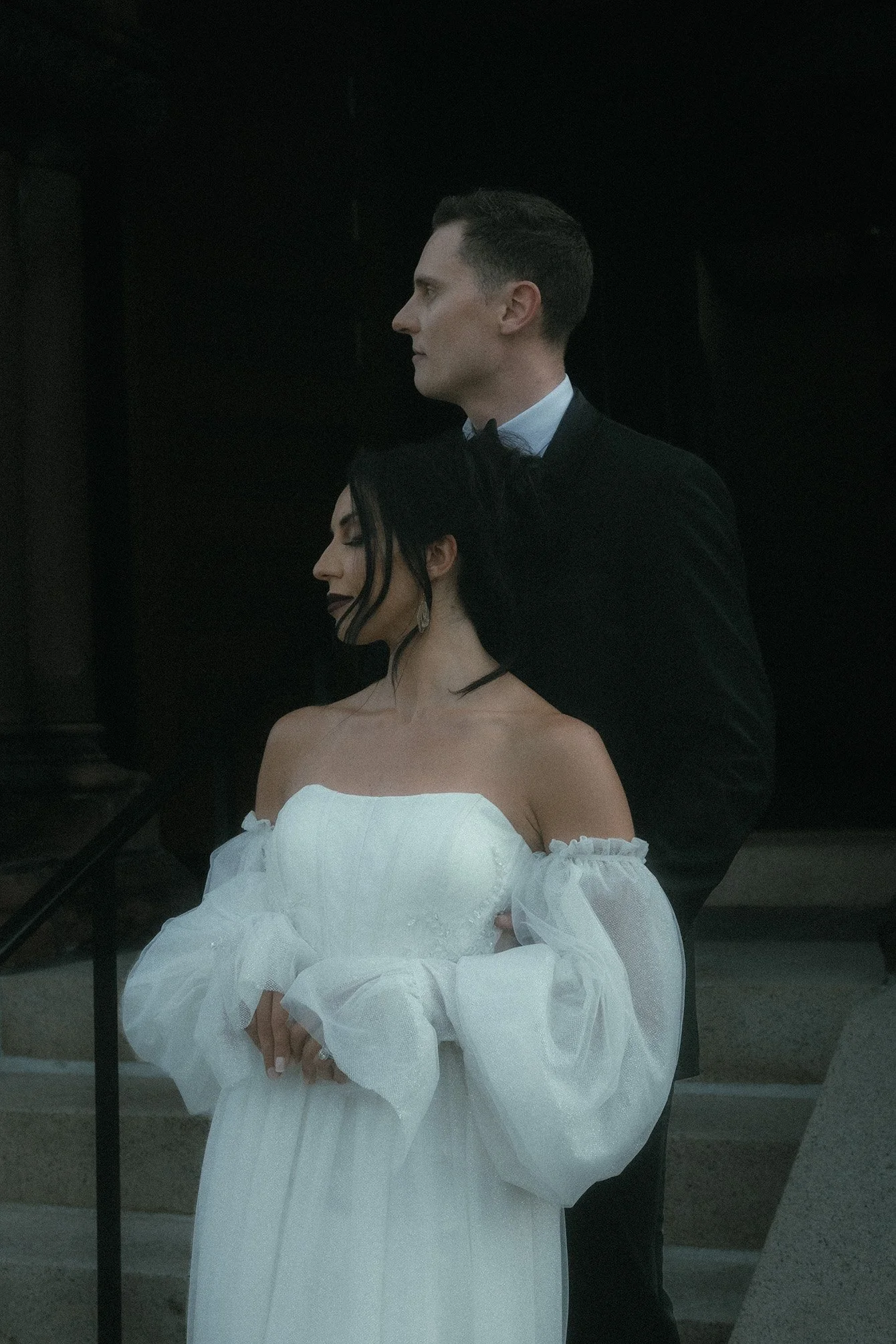 A couple dressed in formal attire, with the woman in a white off-shoulder gown and the man in a black tuxedo, standing outdoors on stone steps at night.