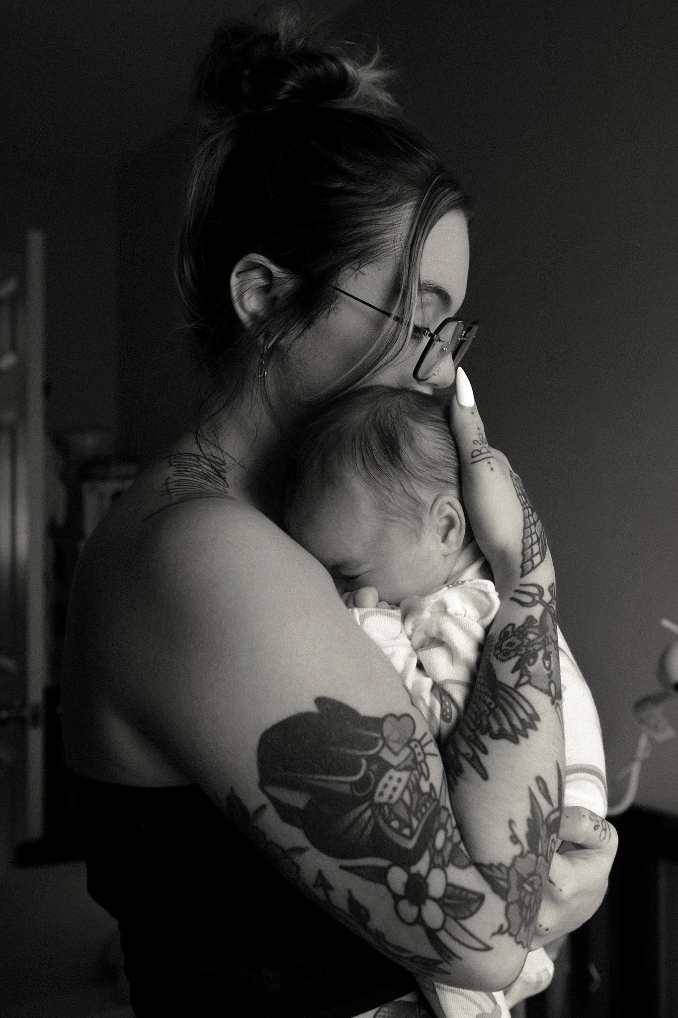 A woman with tattoos and glasses holding a nuzzling, sleeping baby close to her chest, gently touching the baby's head.