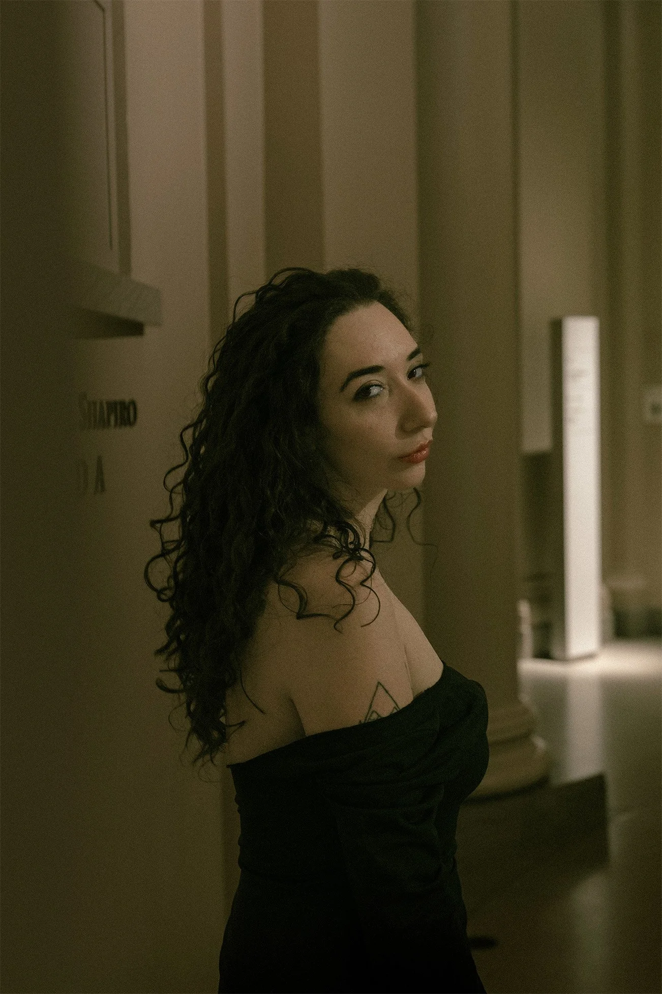 A woman with dark curly hair and makeup, wearing a black off-the-shoulder top, standing in a dimly lit fine art museum hallway, looking over her shoulder at the camera as it peeks from behind the wall.