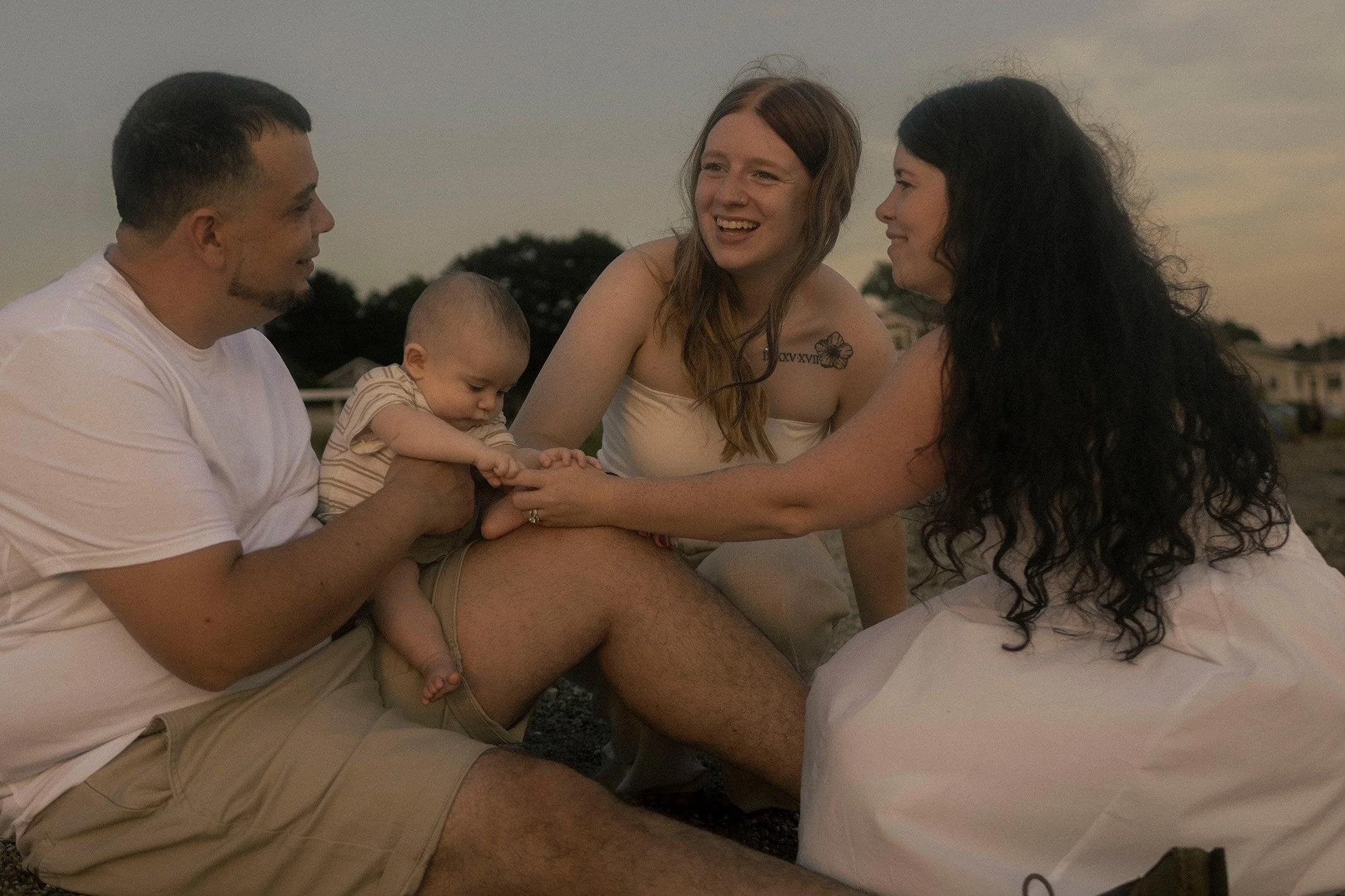 A family of four playing and sitting together outdoors on the beach at sunset, with a man and woman holding a toddler and a young girl touching the man's knee.