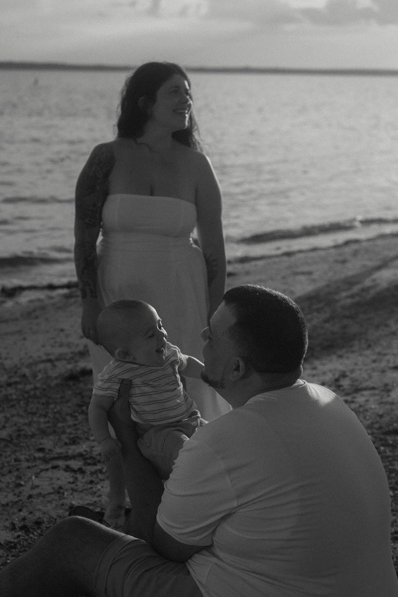A family on the beach during sunset: a woman (the mother) in a strapless dress stands, looking off to the side as she smiles, while a man sits (the father) holding a laughing baby faces each other near the water.