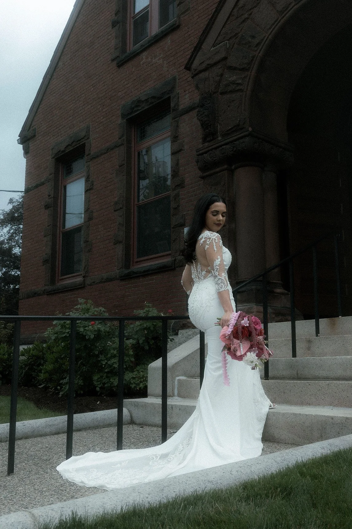 A bride with dark hair wearing a white lace wedding gown holding a bouquet of pink flowers on the steps of a brick building