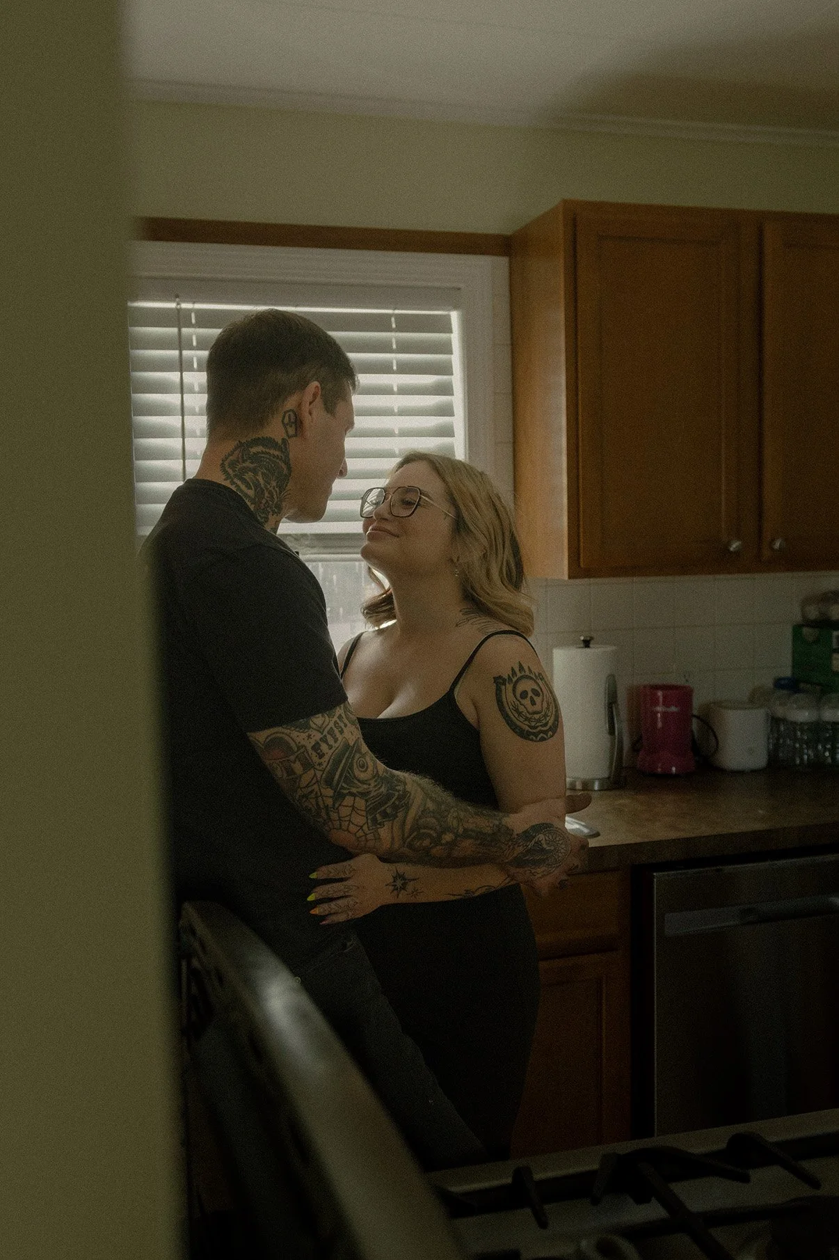 A young man and woman are standing close in a kitchen, looking at each other affectionately. The man has tattoos on his arms and neck, and the woman has a tattoo on her arm and is wearing glasses and a black top.