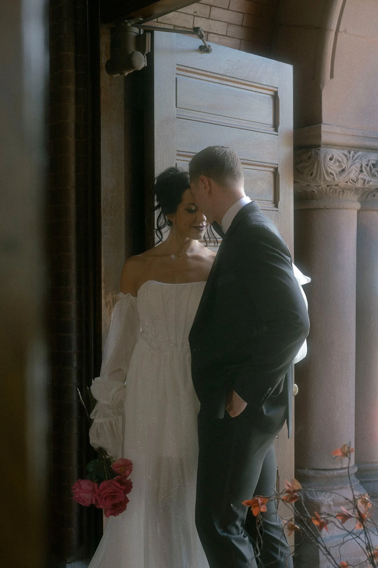 A couple dressed in wedding attire standing close together near a doorway, with the bride holding a bouquet of pink roses.