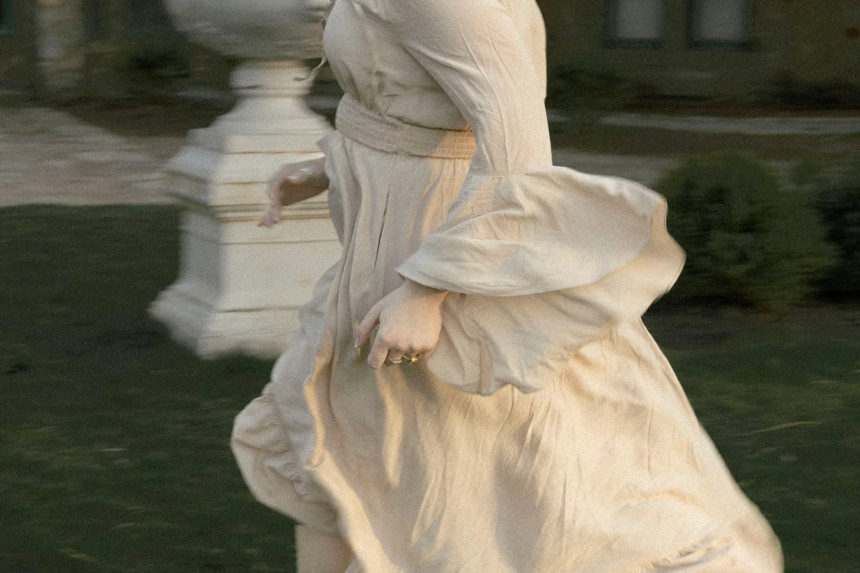 Close-up of a person wearing a flowing, light-colored dress, running outdoors near a stone balustrade, with greenery in the background.