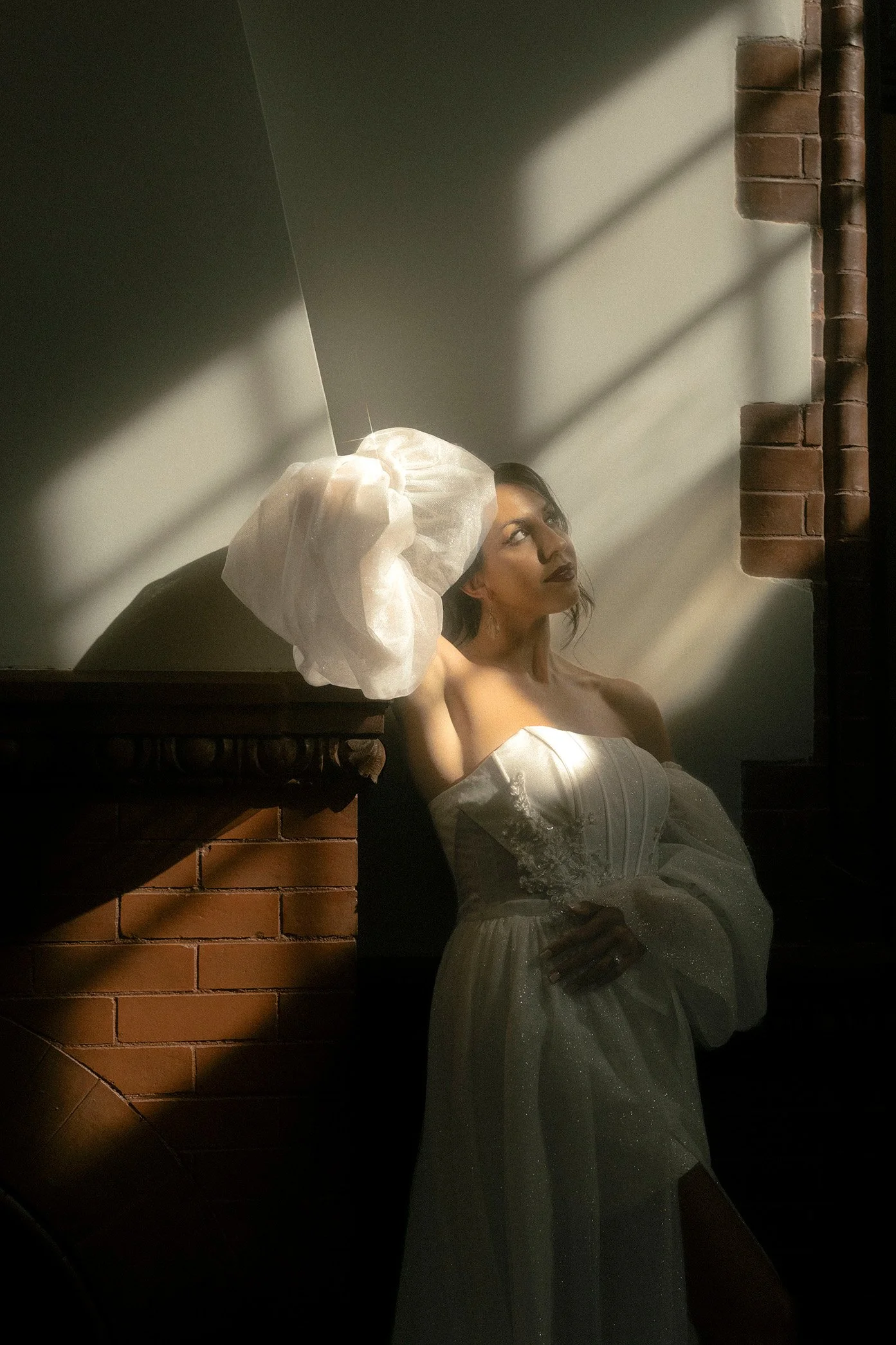 Woman in a strapless white dress with long puffy sleeves, standing against a wall with brick and plain surfaces, illuminated by dramatic sunlight.