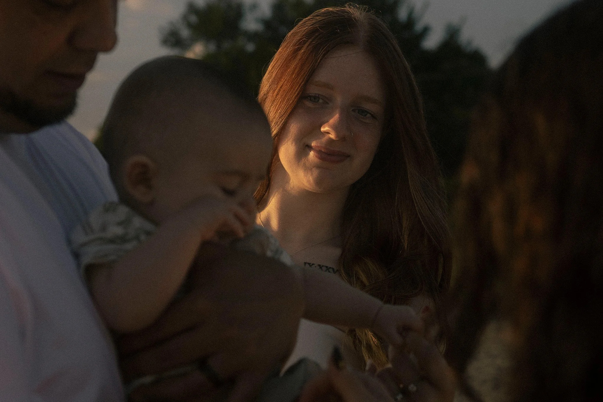 A group of people outdoors during sunset, with a woman with long red hair smiling at a child being held by a man. The background shows trees and a darkening sky.