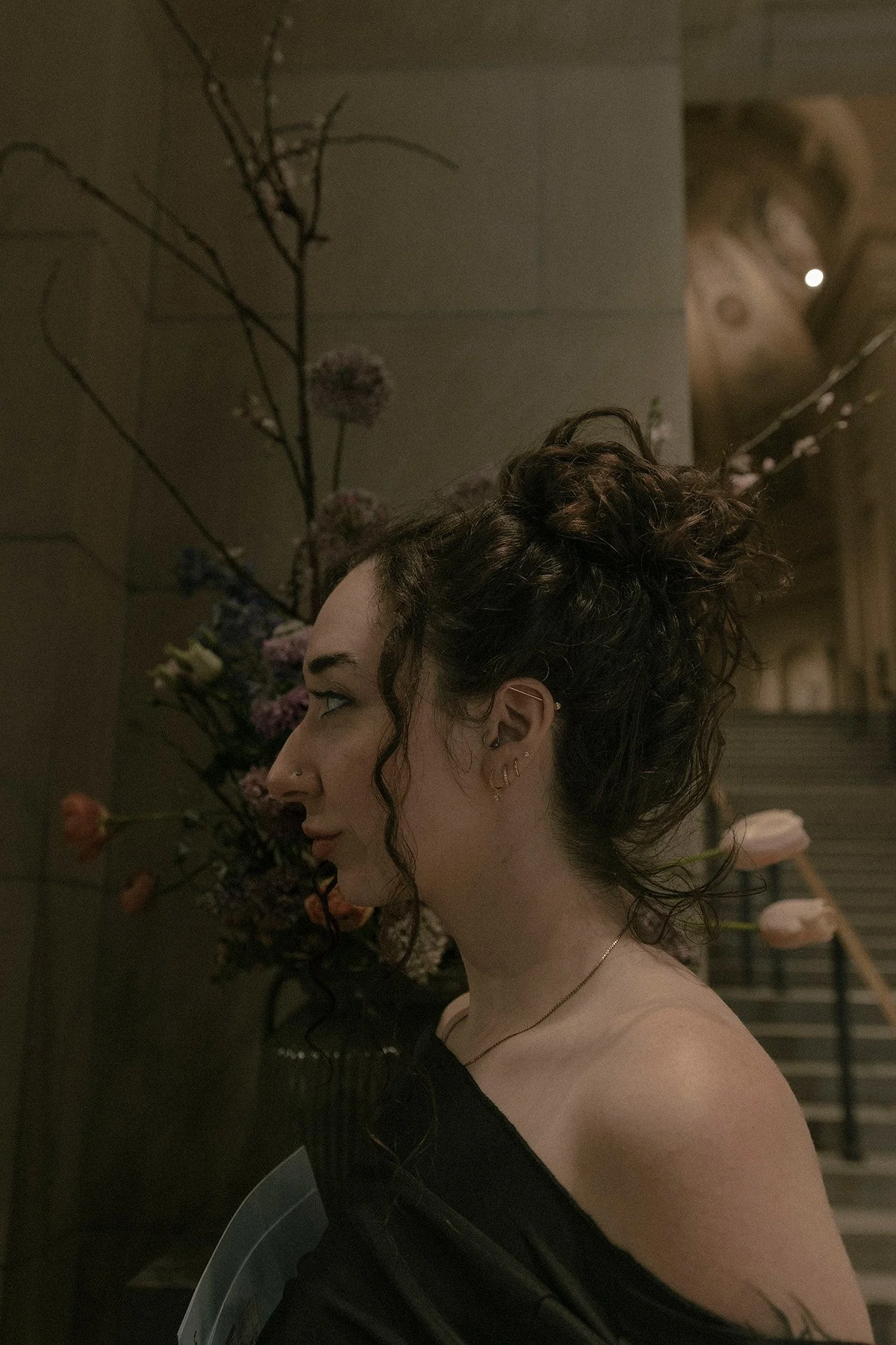 A woman with curly dark hair styled in an updo, multiple earrings, and a nose piercing, standing sideways in front of a floral arrangement and a staircase in a dimly lit space in a fine art museum. 