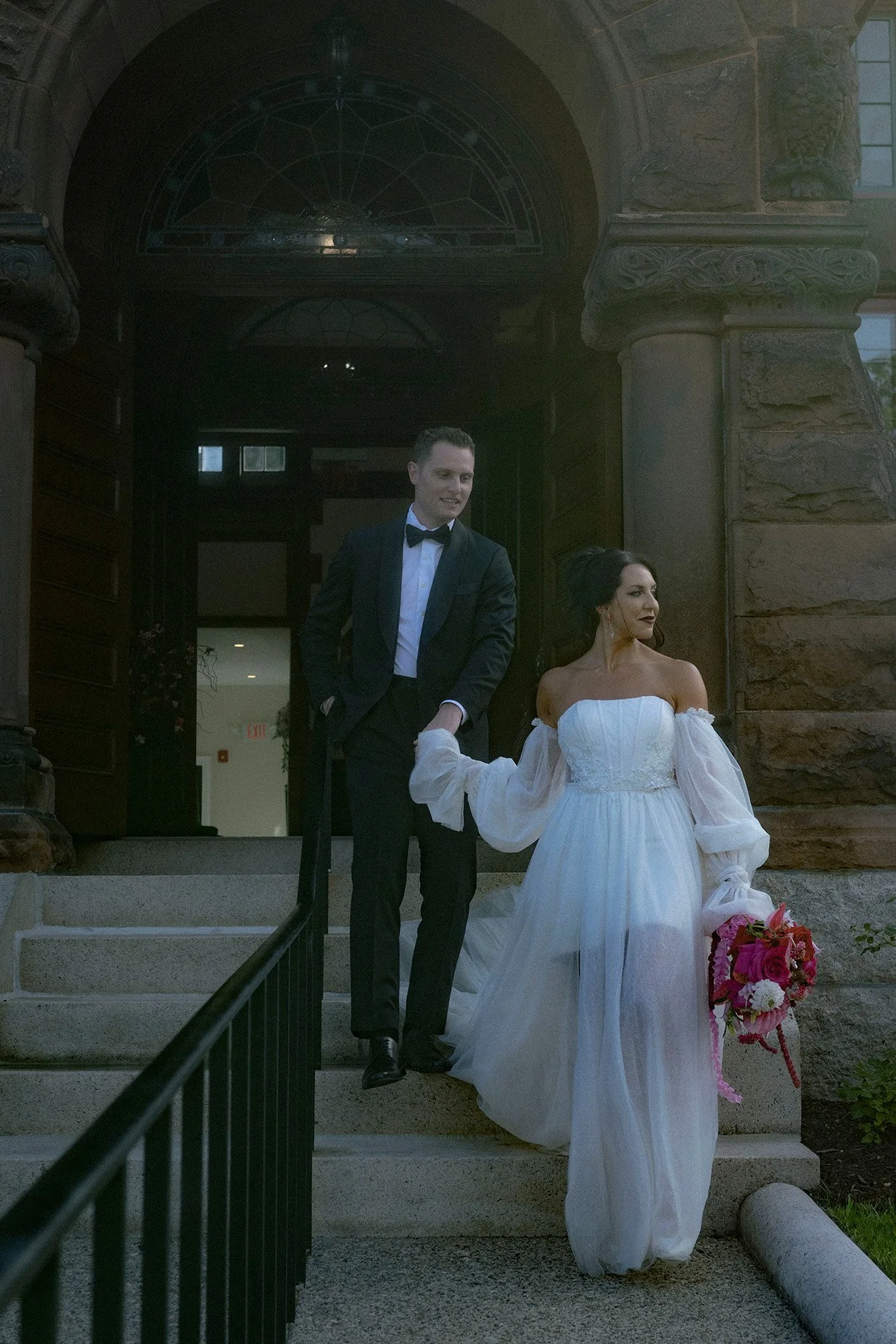 A couple walking down the stairs at a wedding. The woman is in a white wedding dress holding a bouquet, and the man is in a black tuxedo with a bow tie. They look off to the side, smiling, as they walk toward the camera. 