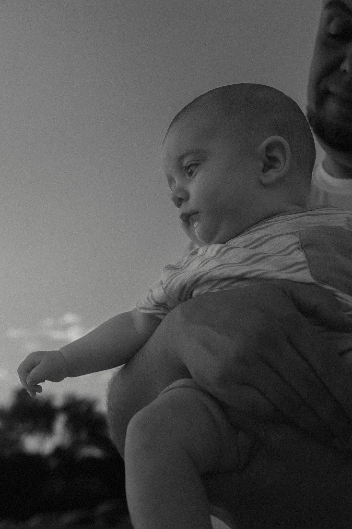 A black and white photo of a young child being held by an adult, with the child looking to the side, outdoors at the beach with a blurred sky and trees in the background.