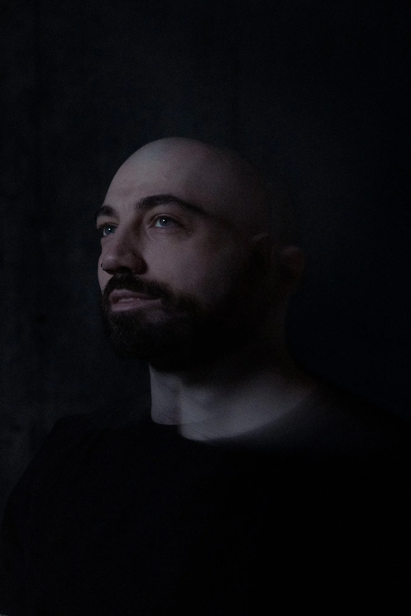 A bald man (James) (Photographer) with a dark brown beard and mustache looking slightly upward, illuminated softly from the front against a dark background. A motion blur affect makes his face appear multiple times against a dark background.