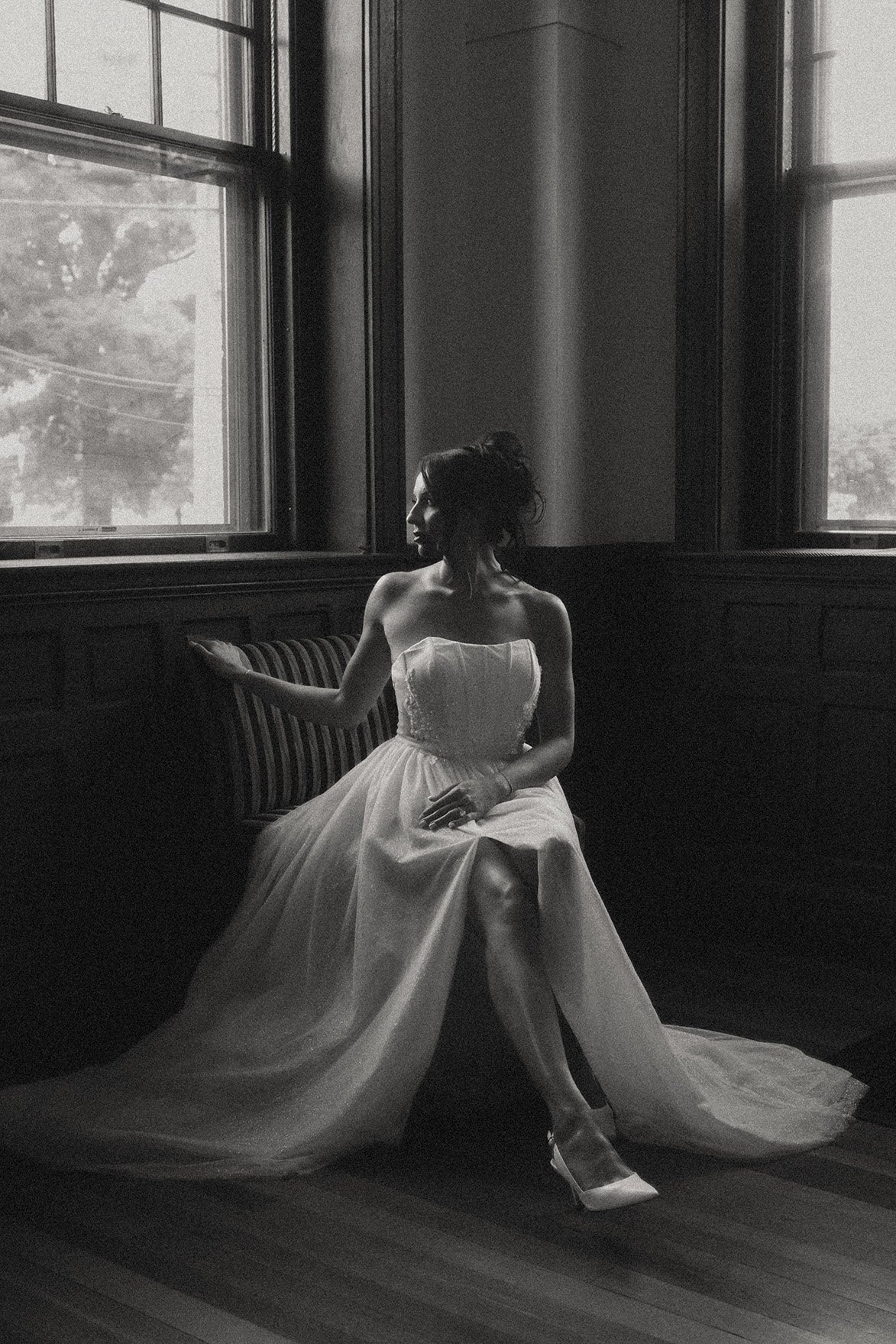 A woman in a strapless wedding dress sitting on a bench near two large windows, with light coming through, black and white photograph.