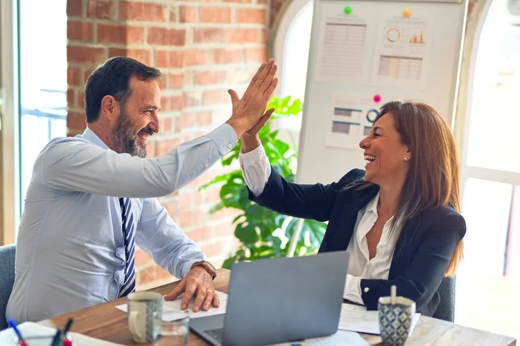 A man and woman in business attire celebrating with a high five at a meeting table in an office