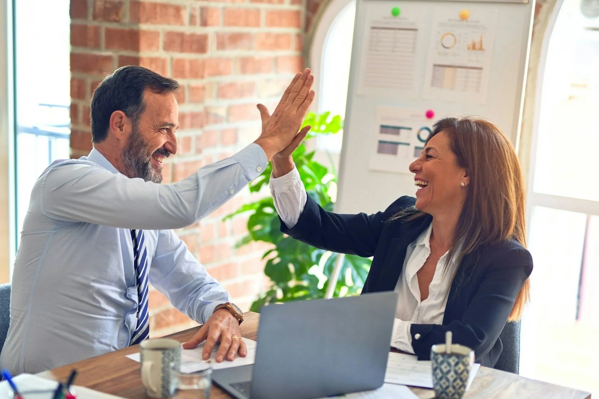 Two colleagues, a man and a woman, give each other a high five in a bright office with a brick wall and chart paper in the background.
