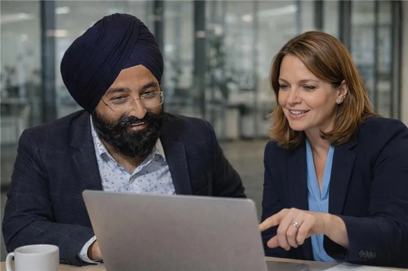 Man wearing turban and glasses and woman with shoulder-length hair looking at a laptop together in a modern office setting.