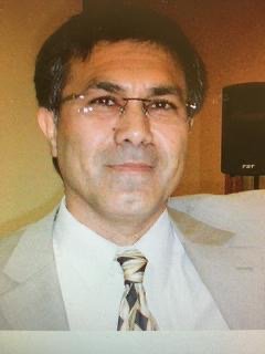 A man wearing glasses, a light-colored suit, and a striped tie, smiling at the camera indoors.