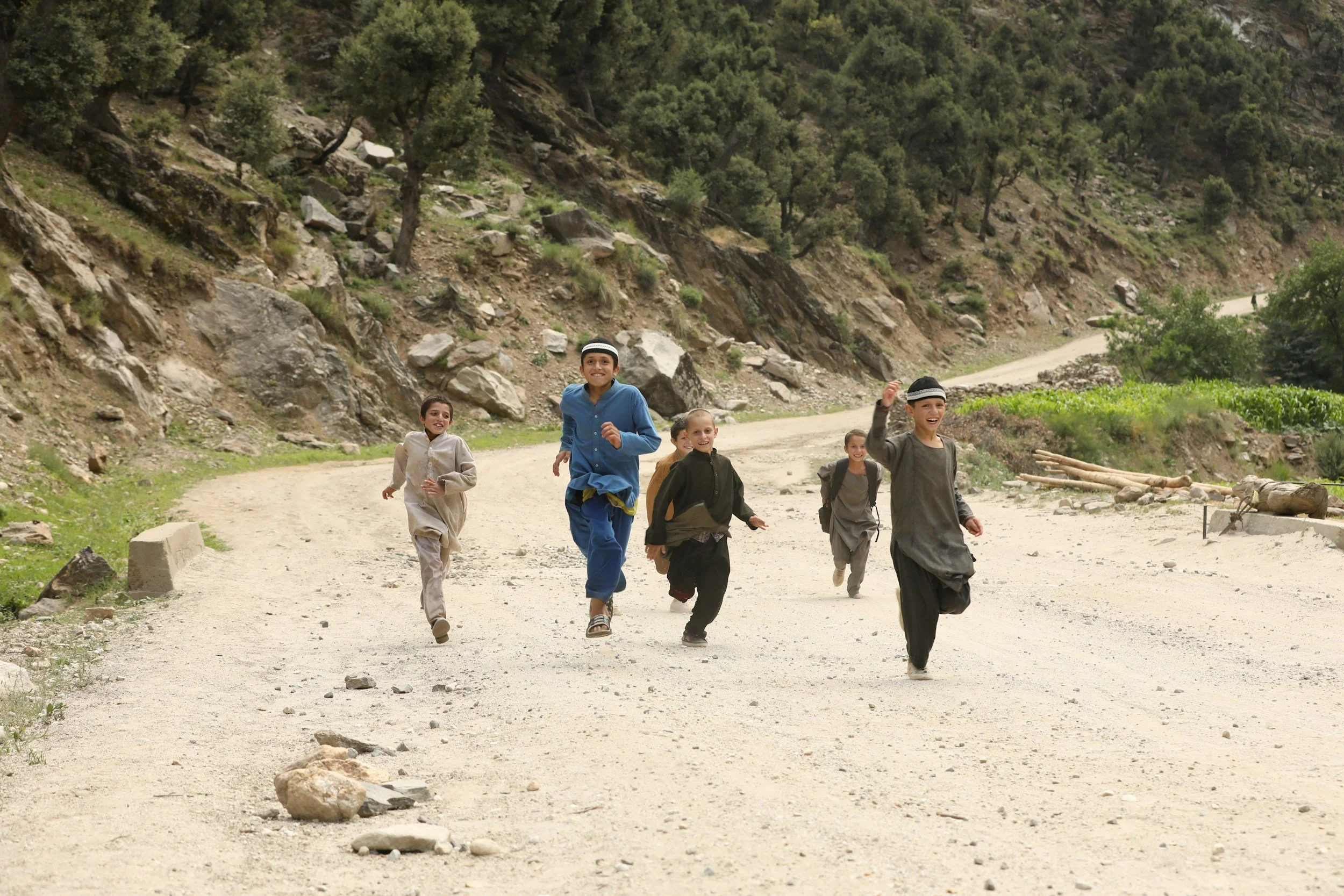 Group of children running and playing on a dirt road in a mountainous area with greenery and rocks.