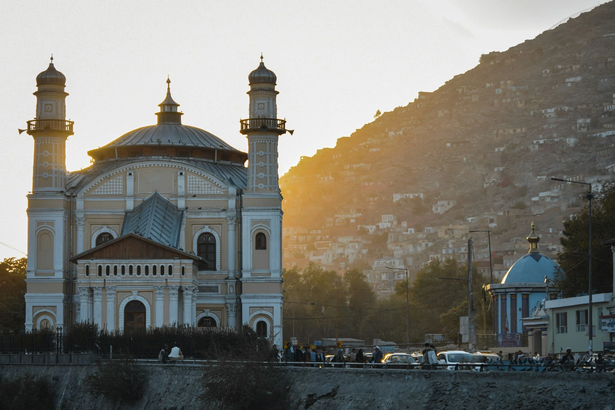 A mosque with two minarets and a domed roof, situated in a mountainous area with a hillside densely packed with small houses, during sunset.