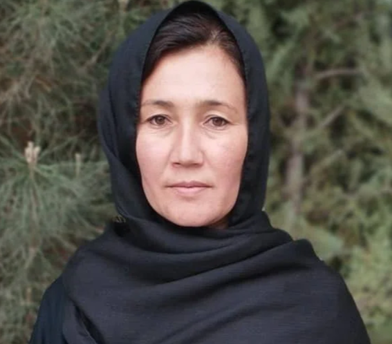 A woman with light skin and dark hair wearing a black headscarf stands outdoors in front of trees.