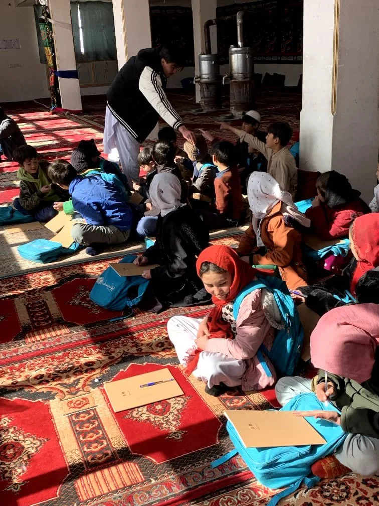 Children sitting on a patterned red carpet in a mosque, participating in a class or activity where a man is handing out items. Some children have backpacks and notebooks, and a few are wearing headscarves.