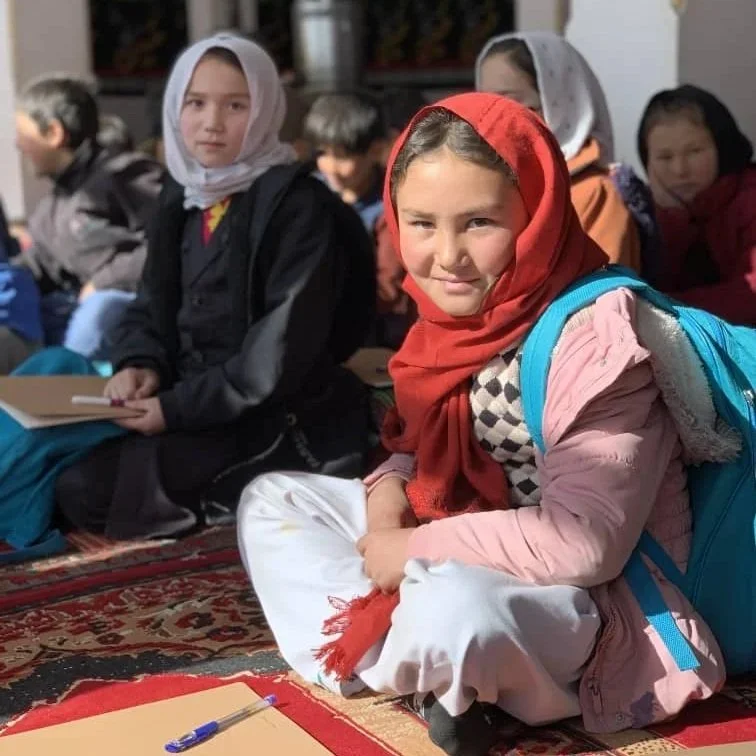 A group of children sitting on a carpet, with a girl in the foreground wearing a red headscarf, pink jacket, and blue backpack, looking at the camera.