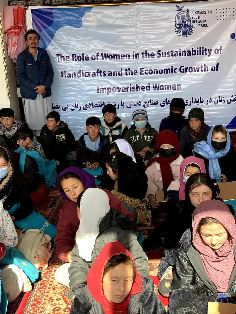 Group of Afghan children and women attending a seminar on women's role in handicrafts and economic growth of impoverished women, with a banner in the background.