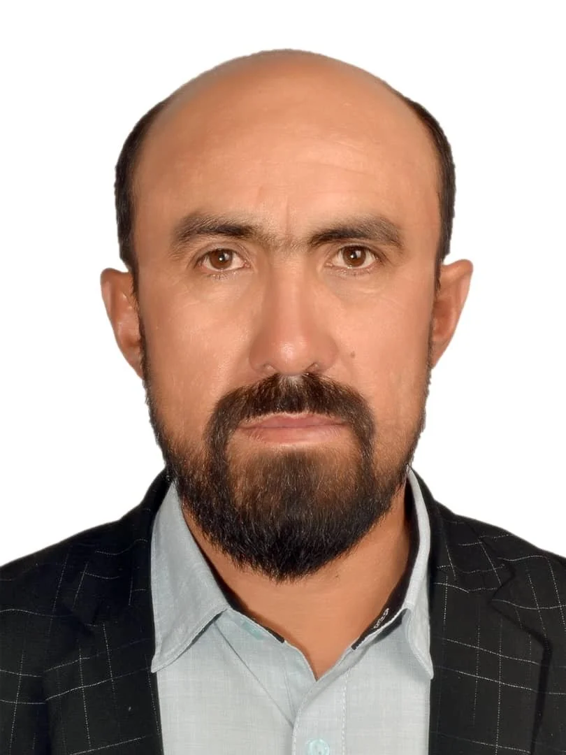 A man with a serious expression, light skin, short dark hair, a full beard, wearing a light blue shirt and a black blazer with white pinstripes, against a plain white background.