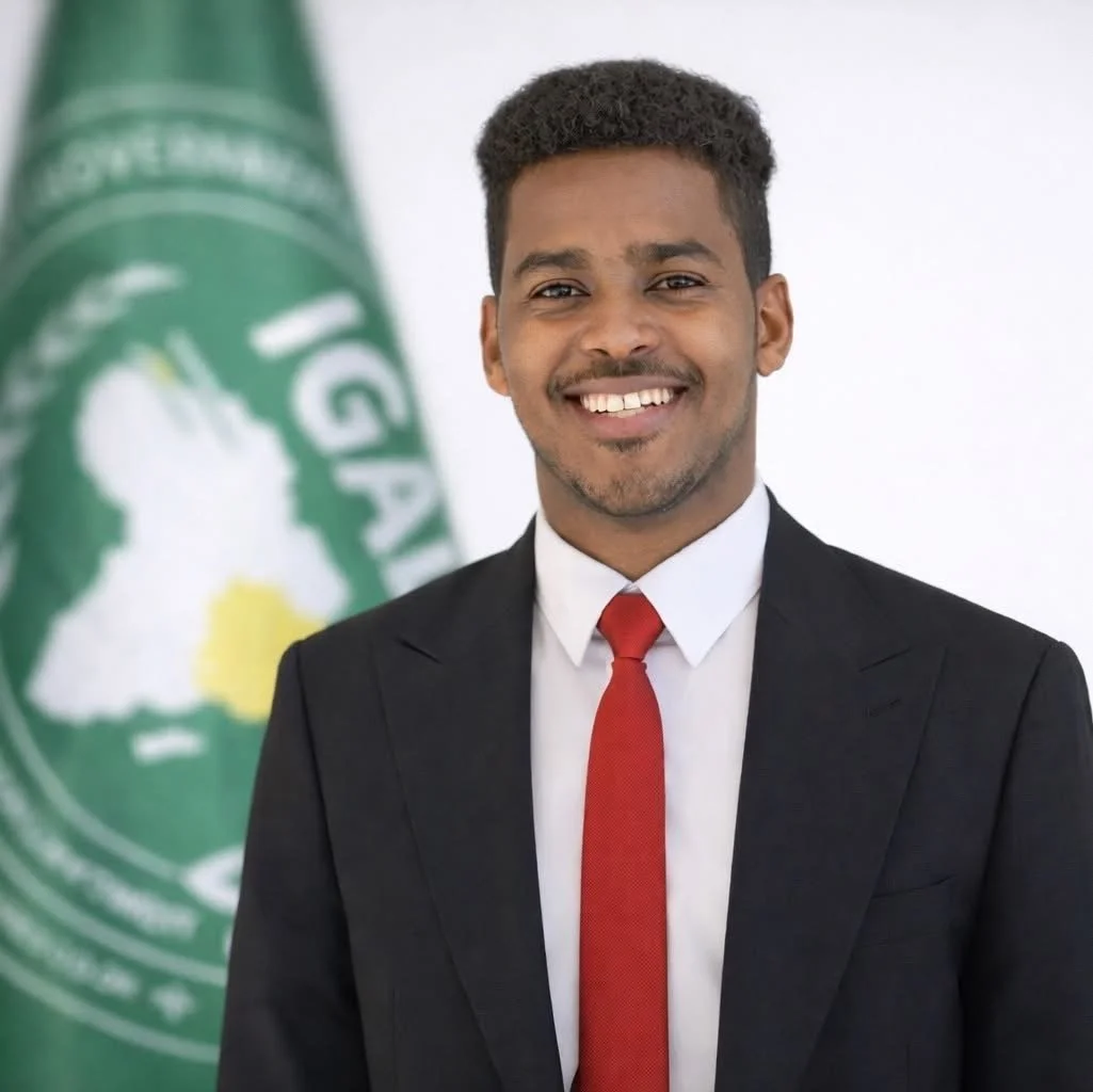 A young man in a suit and red tie smiling, standing in front of a white background with a green and white flag or banner that has an outline of a person and some text.